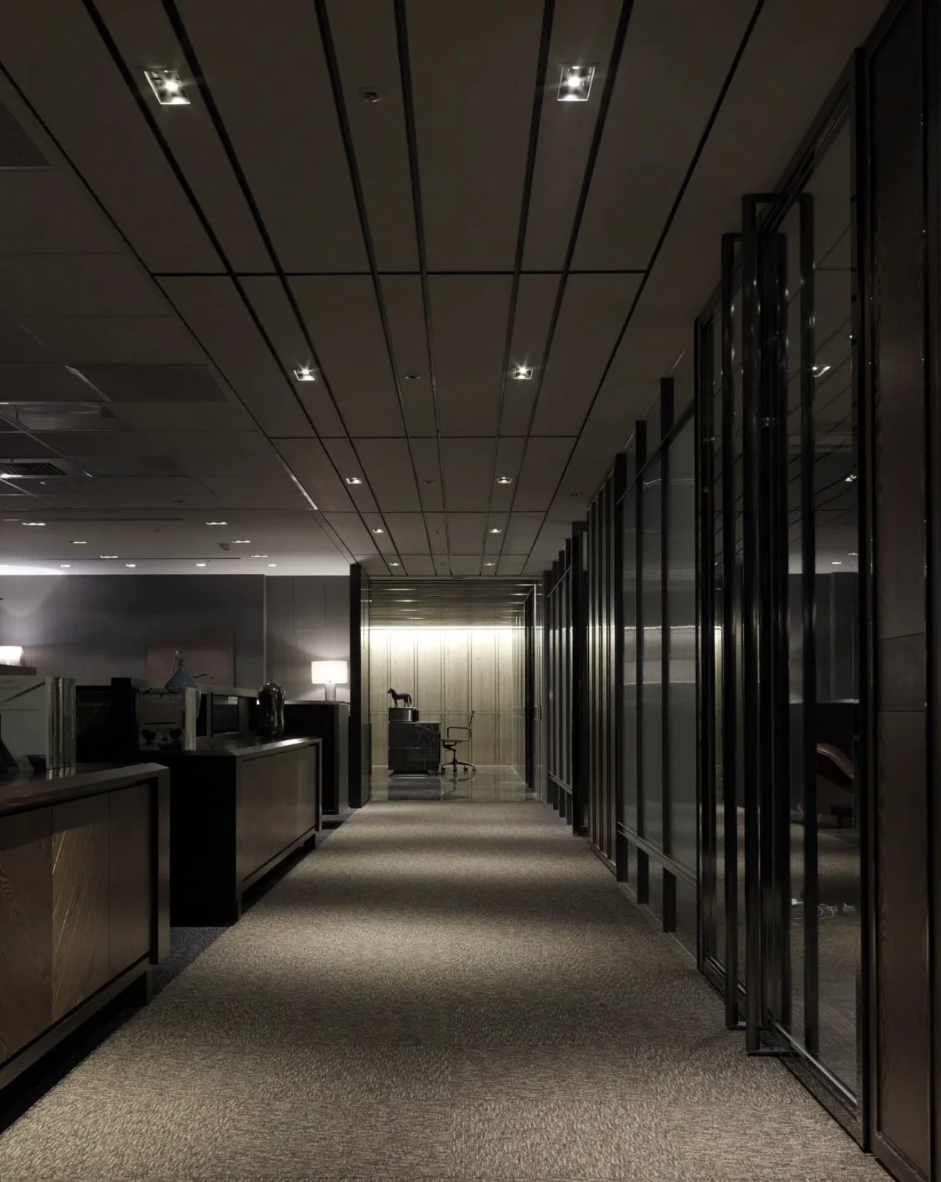 A modern office hallway with dark wood paneling, glass doors, and minimalistic furniture. There are small ceiling lights and a carpeted floor, leading to an area with a desk, chair, and decorative items, including a lamp and a horse figurine.