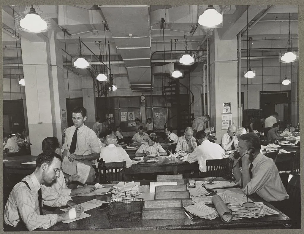 Black and white photo of a busy office filled with people working at desks, some on the phone, some reading or writing, with papers and files scattered across the tables; older style office lighting and a spiral staircase in the background.