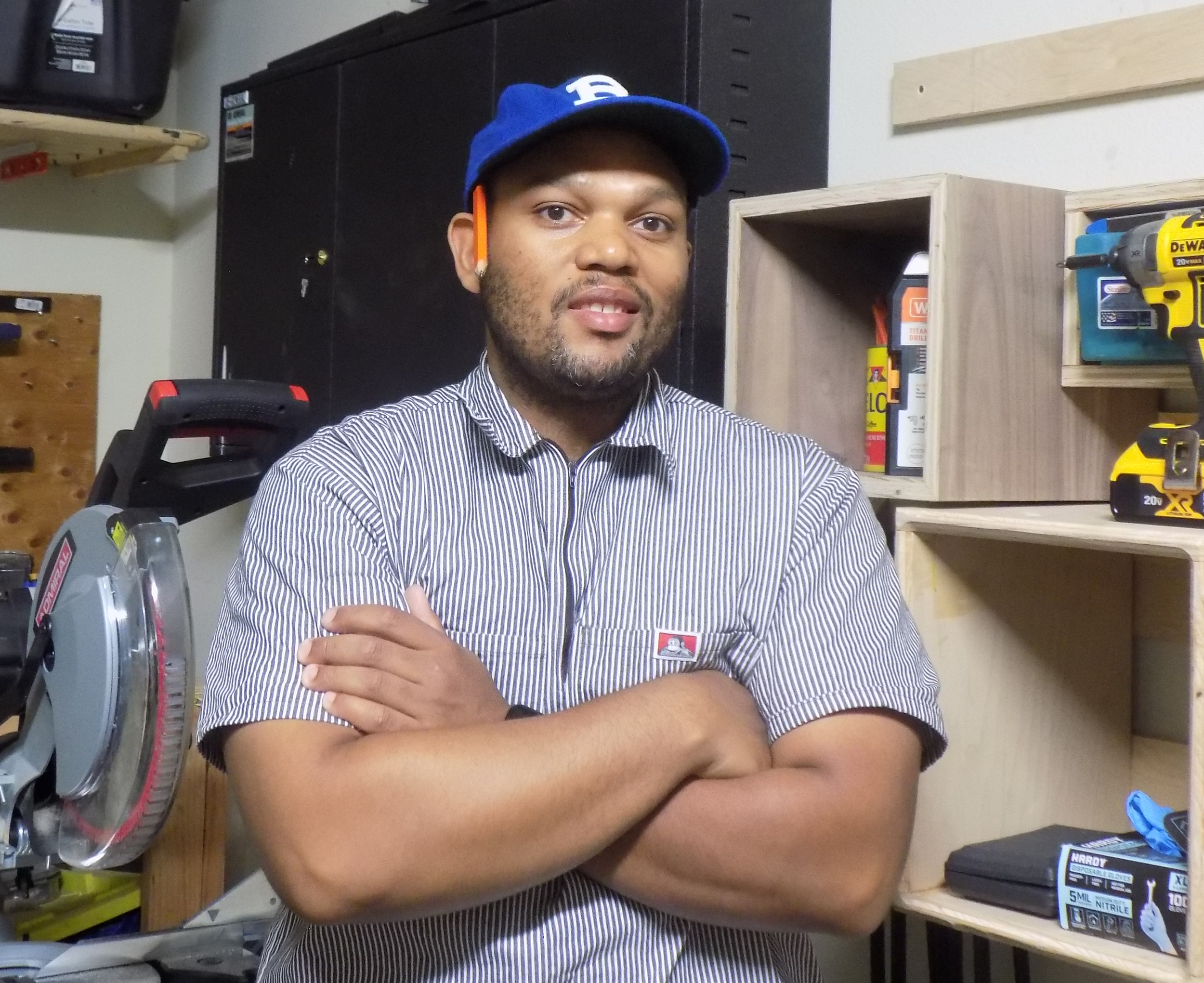 A man with a beard wearing a blue baseball cap and a striped shirt stands with arms crossed in a workshop, surrounded by shelves with tools and equipment.