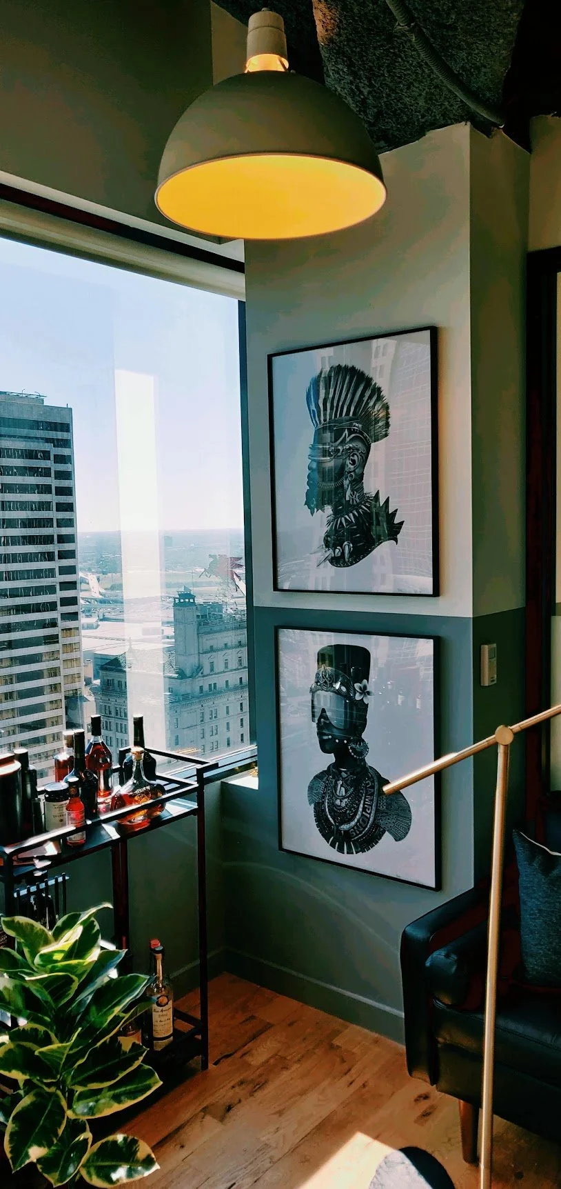 Corner of a modern high-rise apartment with framed African tribal art on wall, large window with city view, bar cart with liquor bottles, green houseplant, and a black armchair near a gold-colored floor lamp.