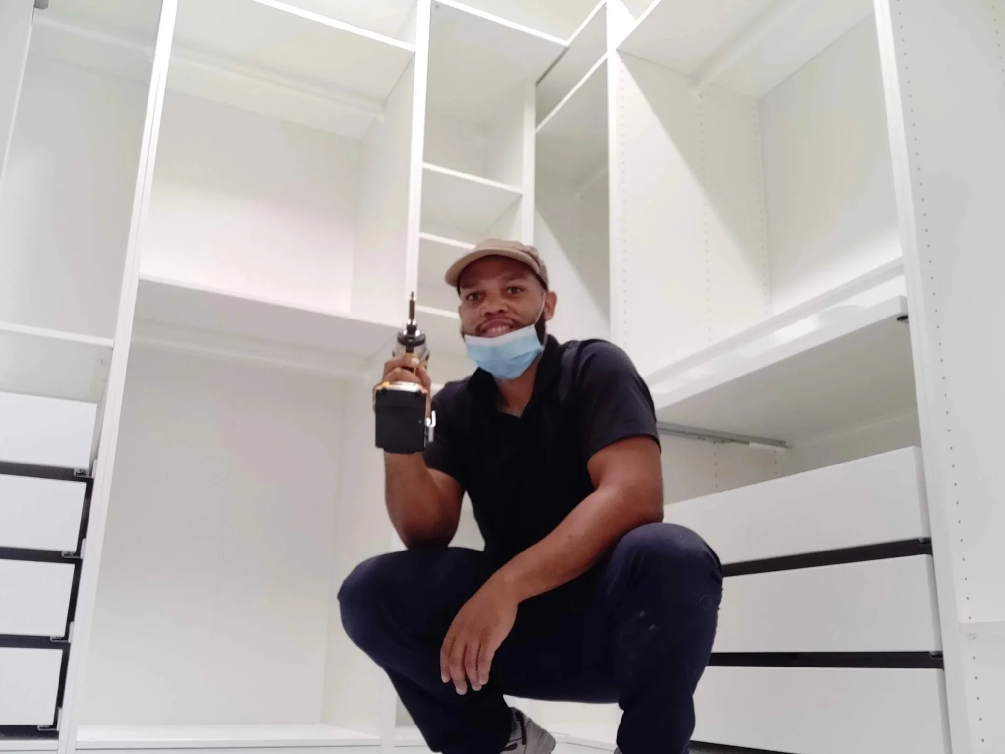 A smiling man wearing a face mask, tan cap, and black shirt, holding a cordless drill, crouching on the floor in front of white shelving units.
