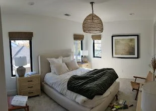 Bedroom with a white bed, beige nightstands, windows with blinds, and a hanging woven light fixture.