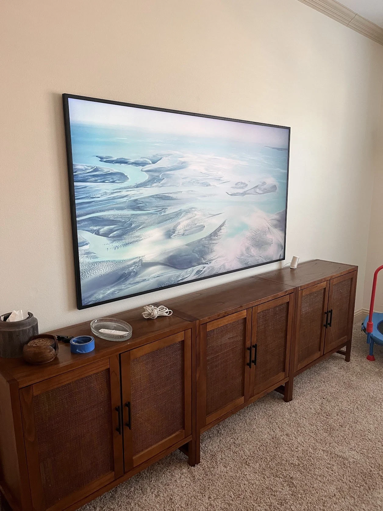 Living room with a large flat-screen TV displaying a snowy landscape, wooden media console with objects including a tissue box, remote control, and decorative items, beige carpet, and part of a red and blue toy or piece of equipment in the corner.