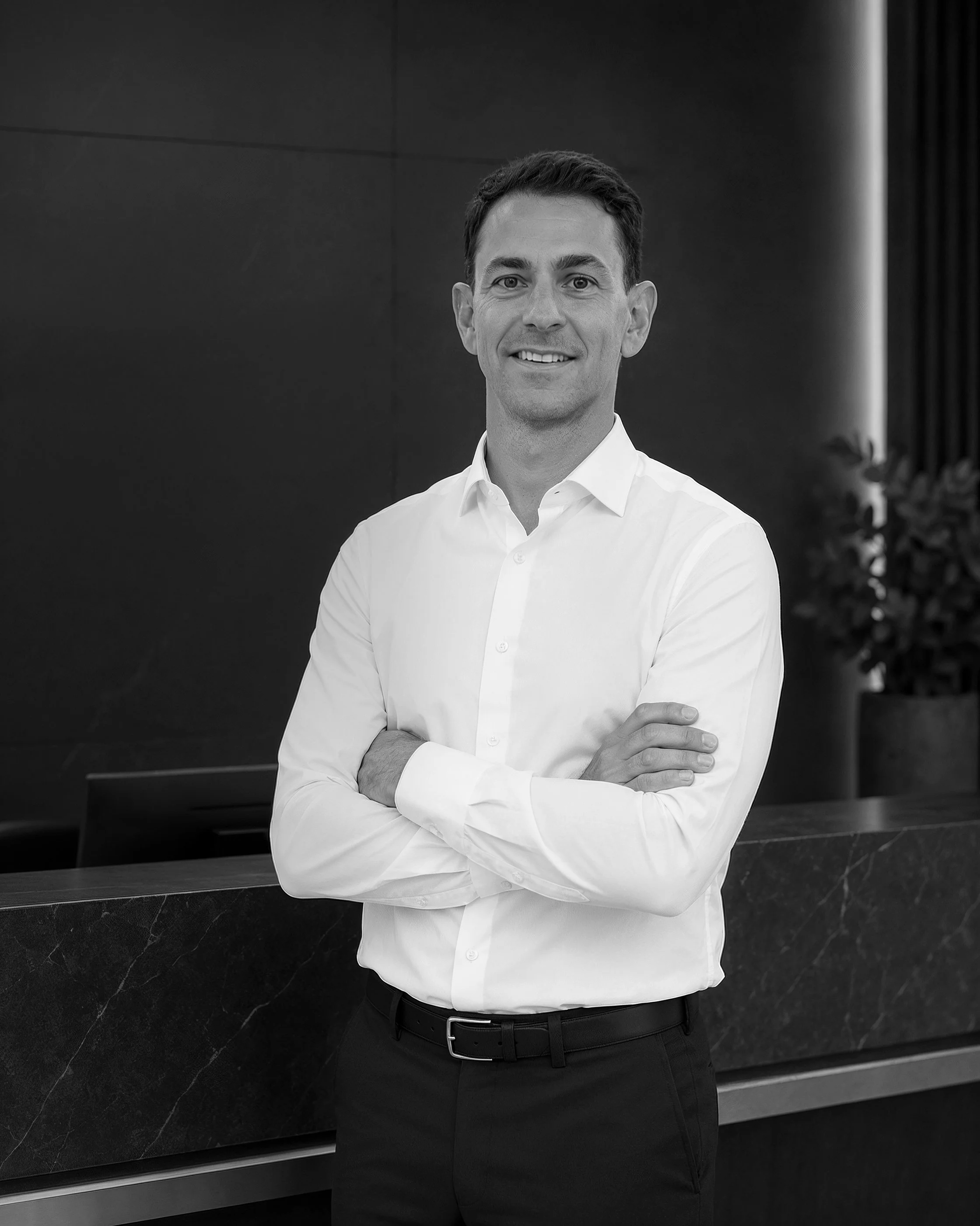 Black and white photo of a man standing with arms crossed in front of a dark wall and a countertop, wearing a white dress shirt and black pants, smiling slightly, facing the camera.