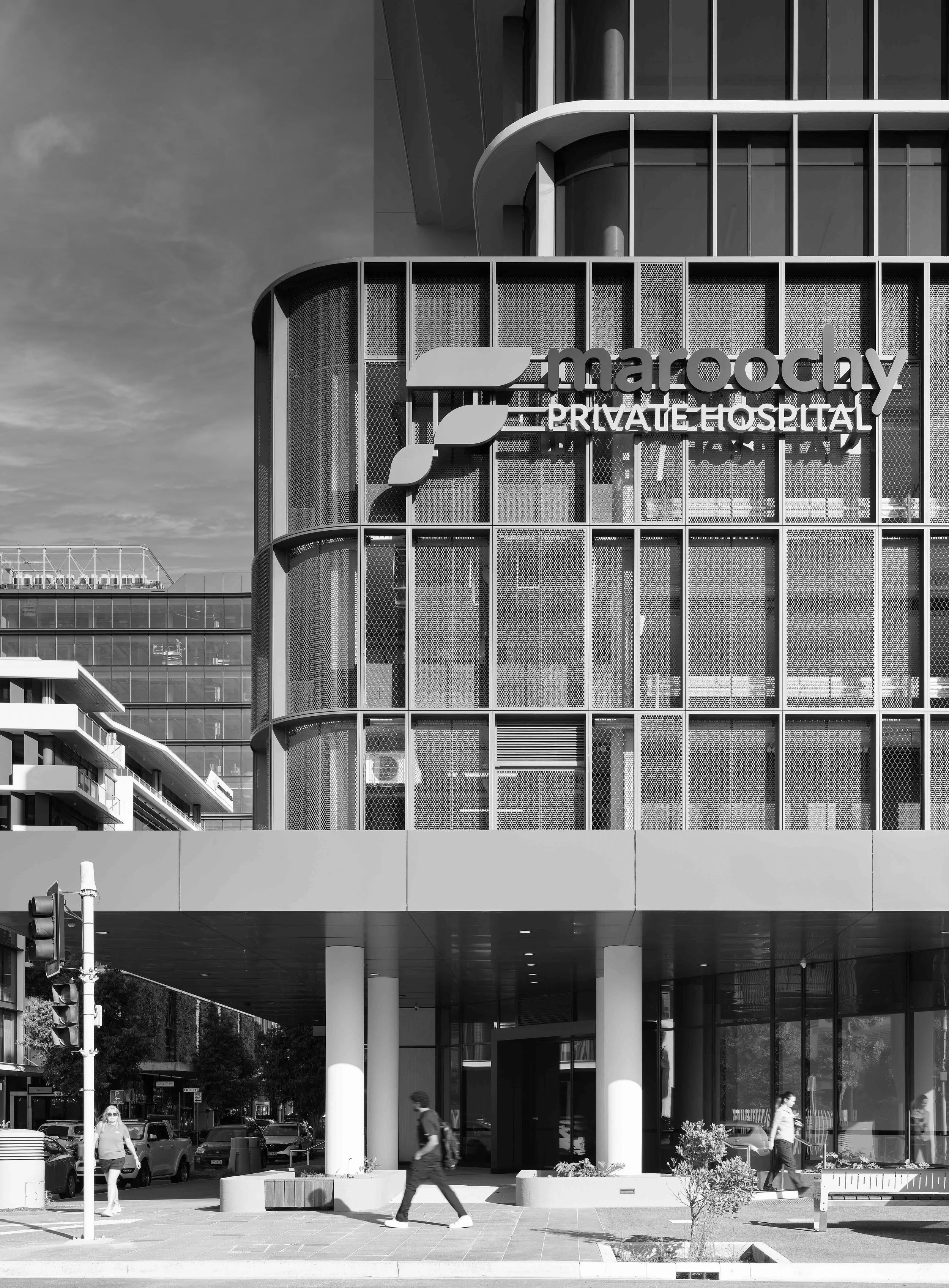Modern multi-story building with sign that reads 'Maroochy Private Hospital', glass facade, and people walking outside in an urban setting.