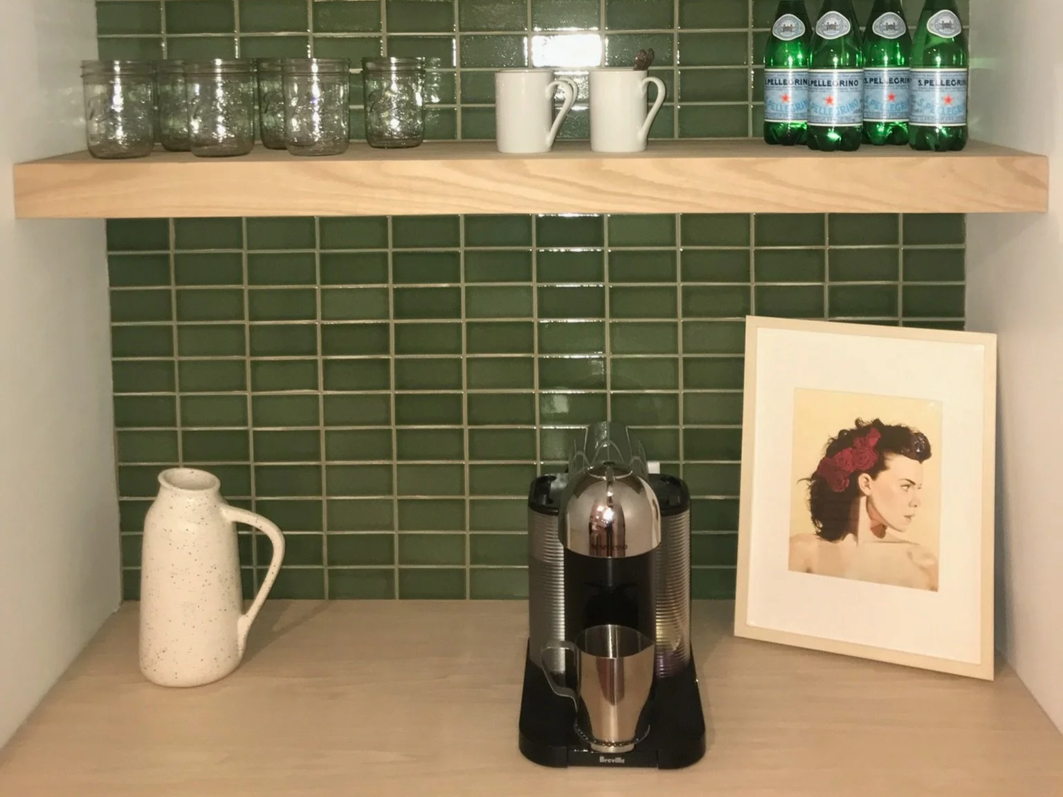 Shelf with glass jars, white mugs, bottles of San Pellegrino sparkling water, a framed picture of a woman with flowers in her hair, and a coffee machine in a kitchen with green tiled wall.
