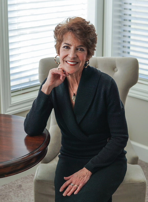 A woman with short, curly brown hair, wearing a black dress with a draped neckline, sitting in a beige chair in front of windows with white blinds, smiling and resting her chin on her hand.