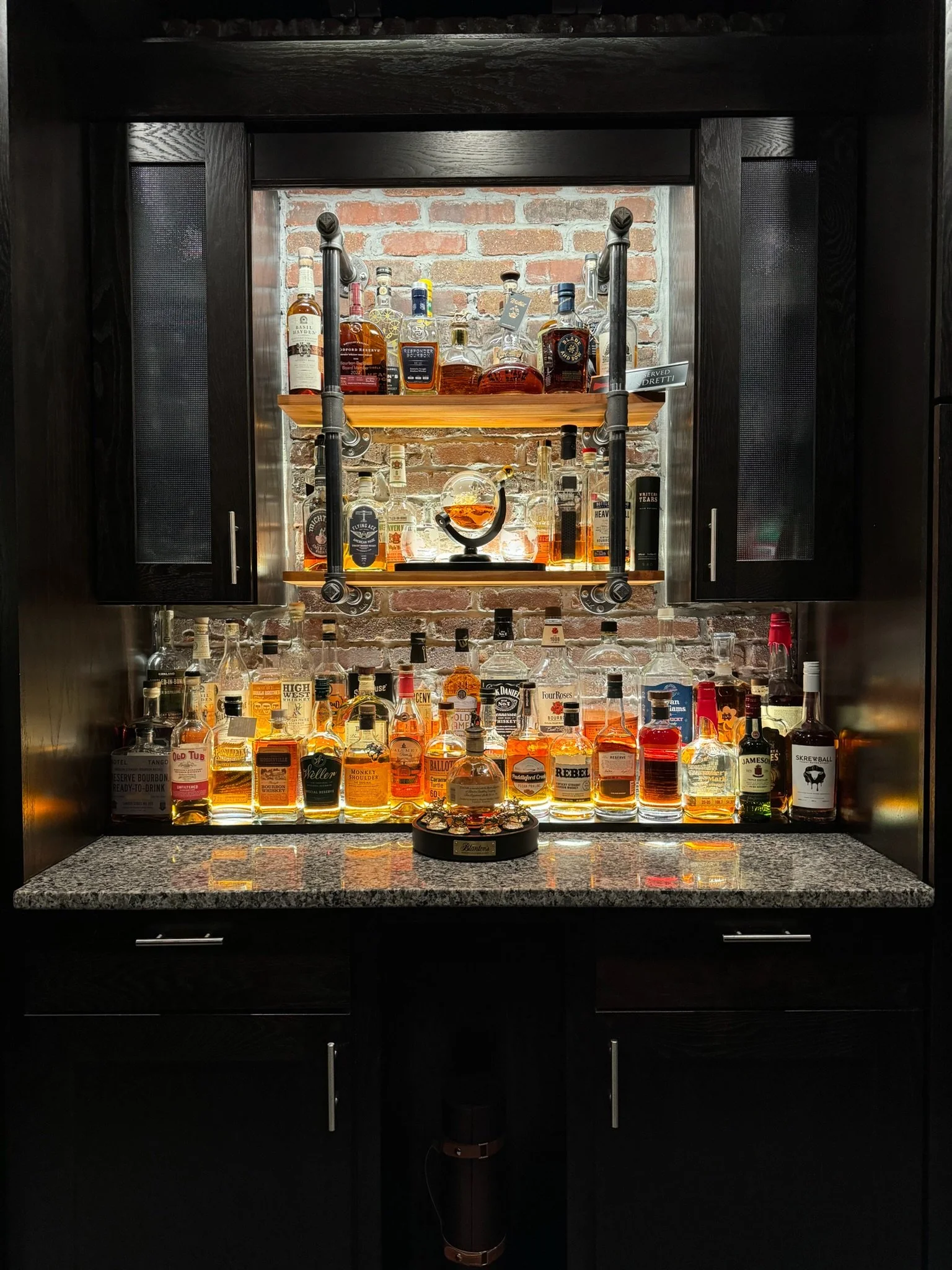 A black cabinet with open doors revealing a collection of liquor bottles, a surgical lamp, and some jewelry on a table.