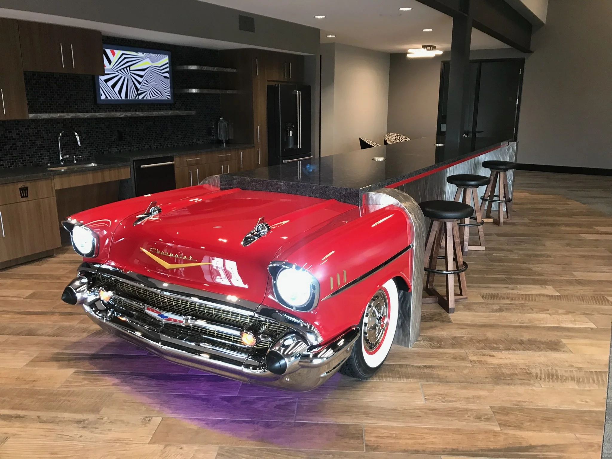 A red vintage Chevrolet Camaro toy car in a modern kitchen with wooden flooring, dark cabinetry, a black countertop, a black refrigerator, and bar stools.