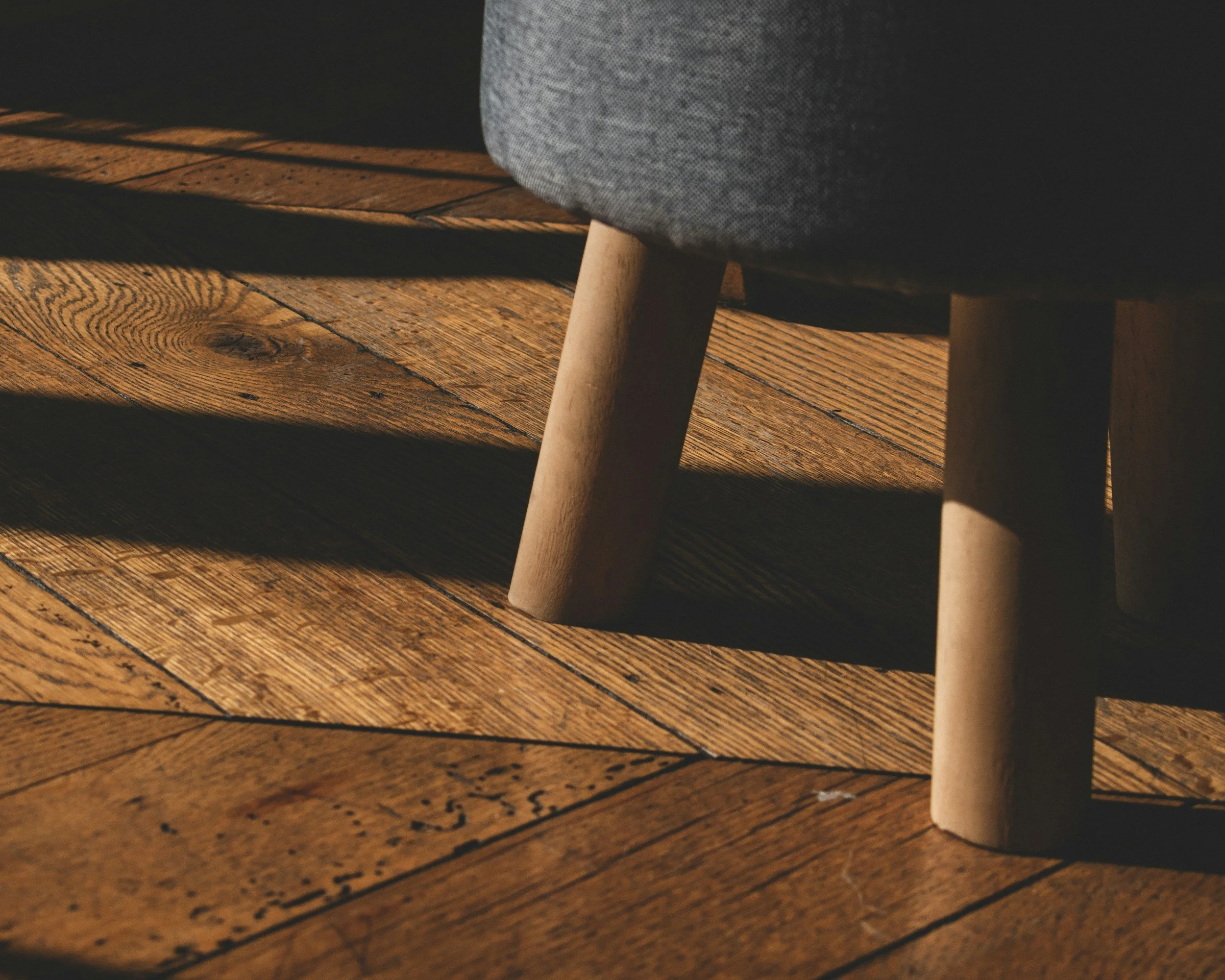 Close-up of a wooden table with a gray upholstered chair partially visible on the right, sunlight casting shadows on the floor.