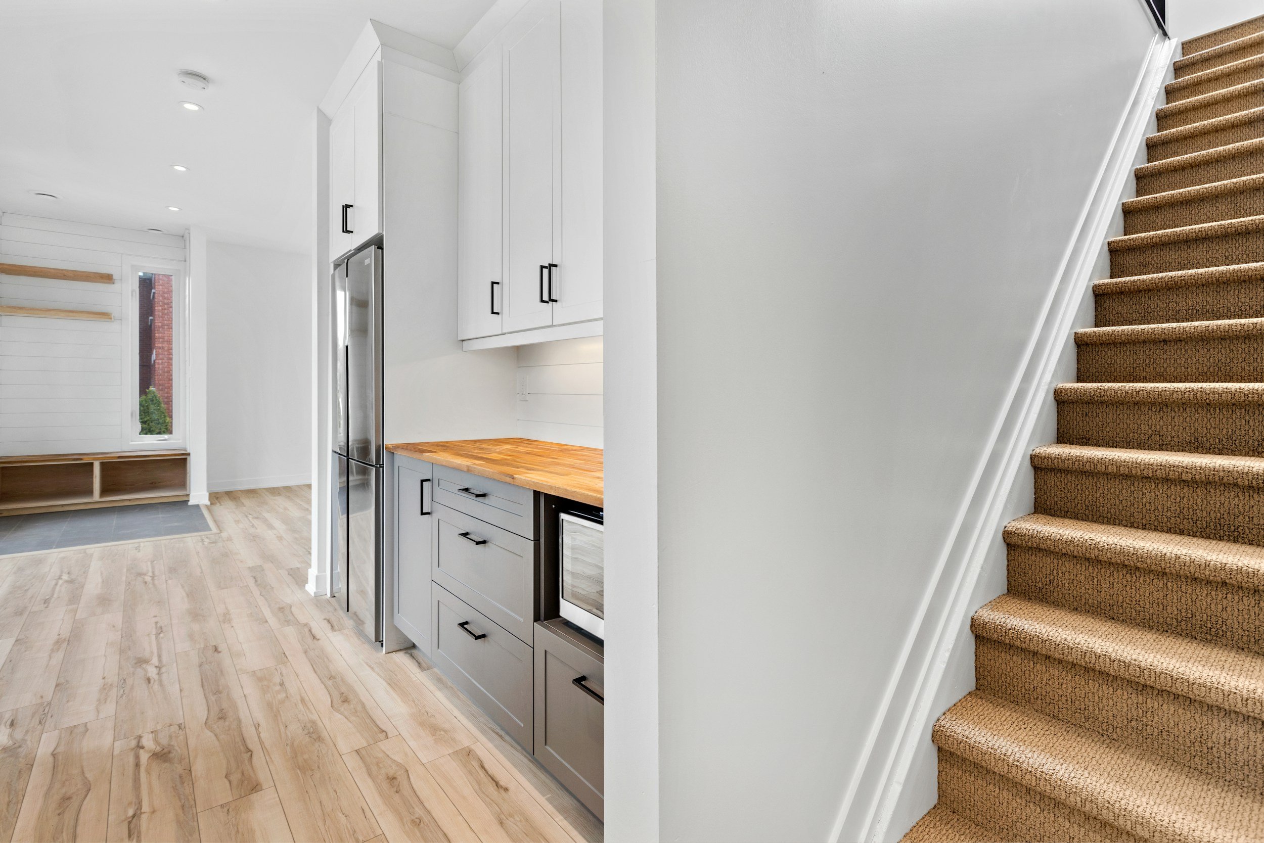 Interior view of a modern home with a staircase, white kitchen cabinets, a wooden countertop, and a living area with wood flooring and a window.