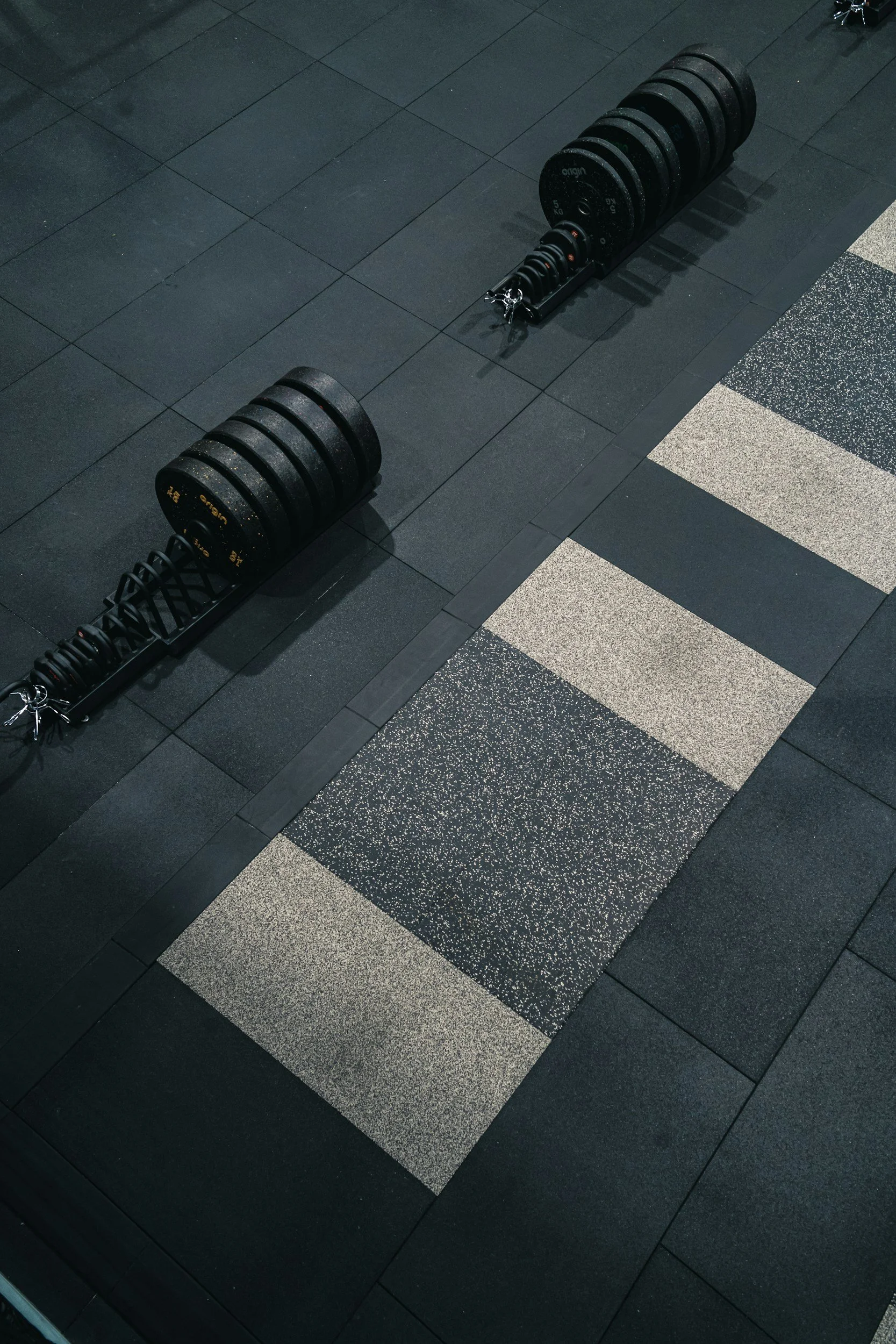 A set of black weight plates on a weight rack on a black gym floor, with part of a patterned grey and black area visible in the corner.