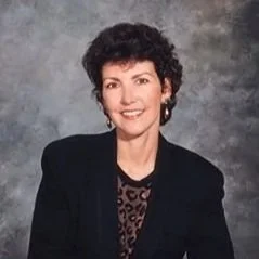 A woman with short curly dark hair wearing a black blazer over a leopard-print top, smiling against a gray textured background.