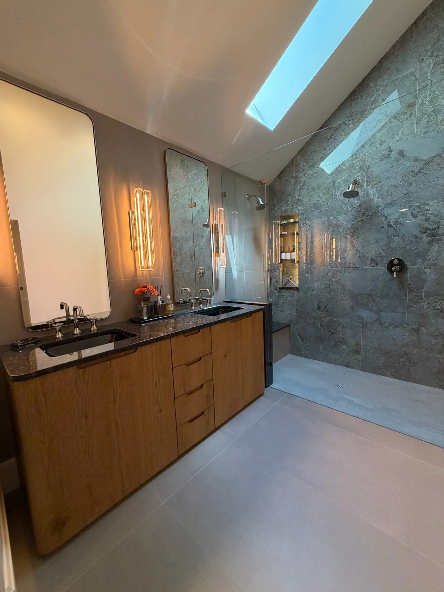Modern bathroom with dual sinks, mirror, wall-mounted lights, wood cabinet, and a walk-in shower with gray marble tiles and a skylight.