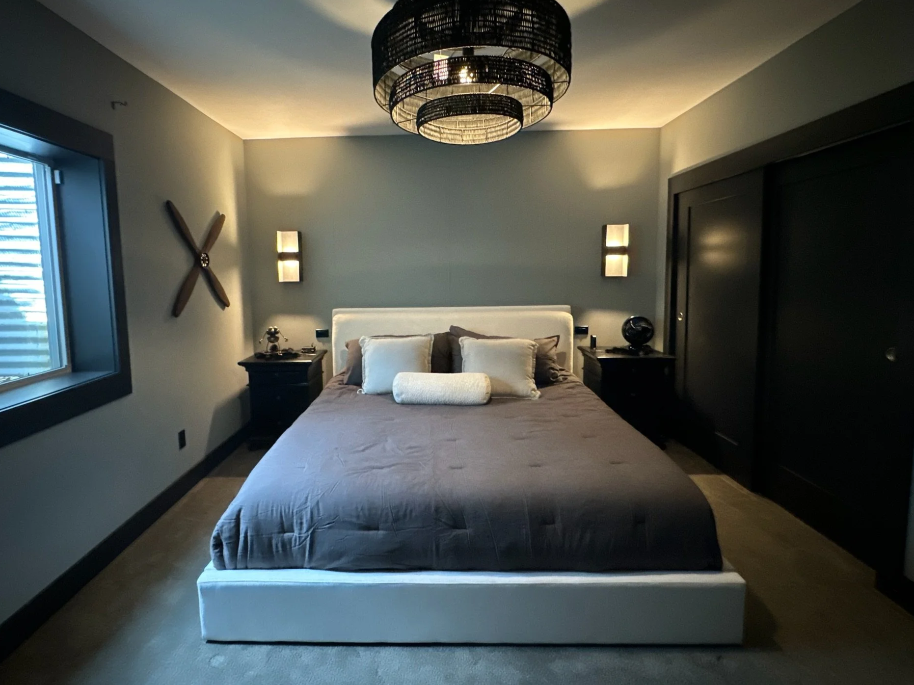 A modern bedroom with a large bed, decorative pillows, and a rolled white pillow, flanked by two dark nightstands with lamps. Artistic propellers are mounted on the light blue wall, and a black circular pendant light hangs from the ceiling. A window with blinds is on the left side.