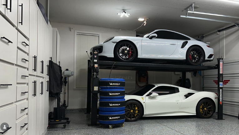 A garage with two white sports cars, one on a lift and one on the ground, surrounded by garage tools and equipment.