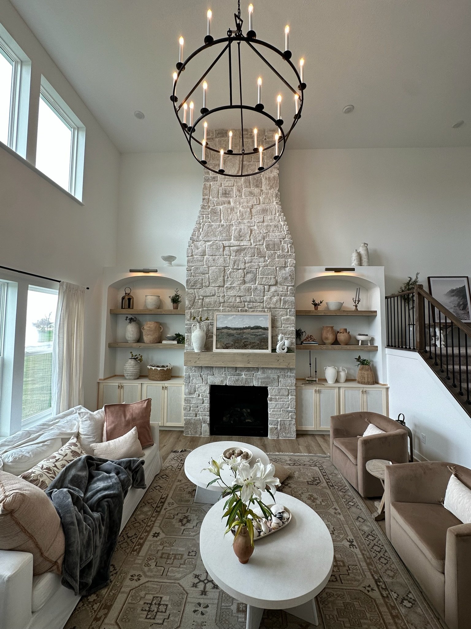 Living room with a stone fireplace, modern decor, beige sofas, and a white oval coffee table with a flower arrangement. Large windows and a chandelier with candles hang from the ceiling.