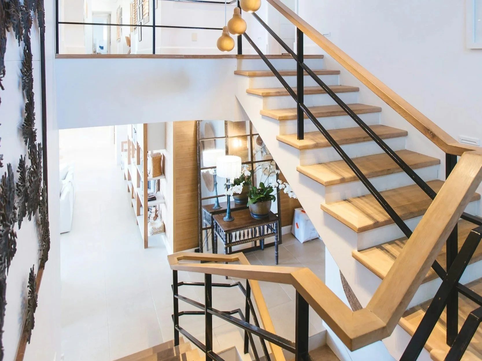Interior view of a modern home featuring a staircase with wooden steps and black metal railings, a glass railing enclosure on the upper level, and a small table with a potted plant and decorative items beneath the staircase. The space is decorated with warm lighting and neutral colors.