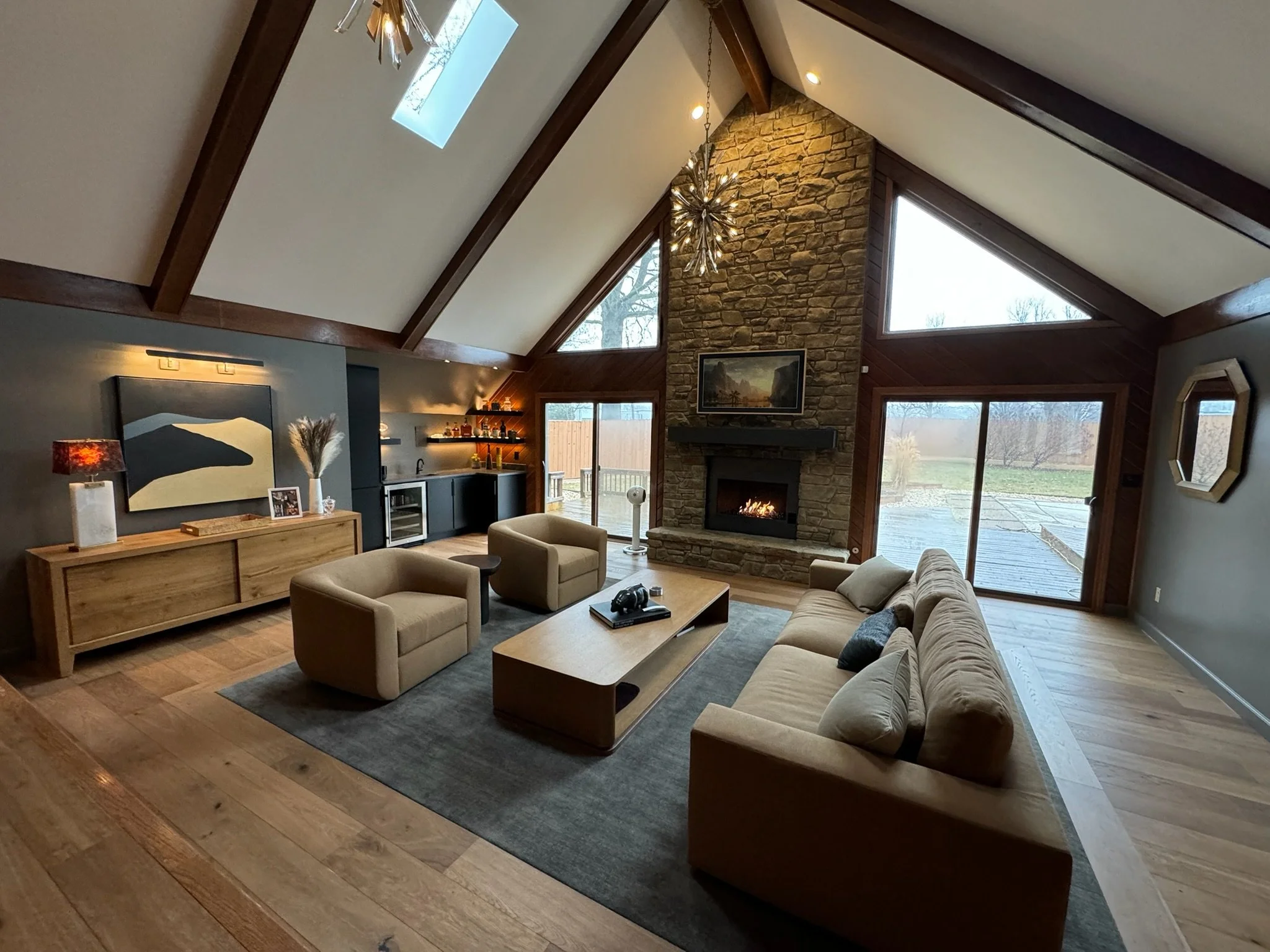 Living room with a sloped ceiling and large windows, featuring a stone fireplace, a chandelier, a beige sofa, armchairs, a wooden coffee table, and a dark cabinet in the corner.