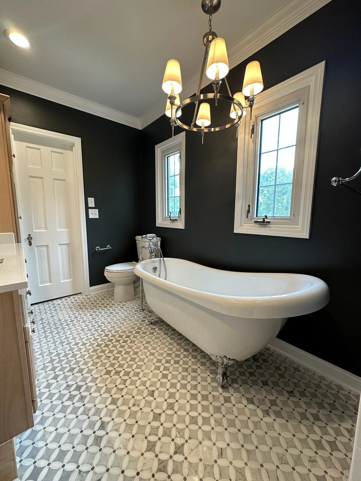 A bathroom with a freestanding clawfoot bathtub, dark blue walls, two windows, a chandelier, a toilet, and a patterned tile floor.