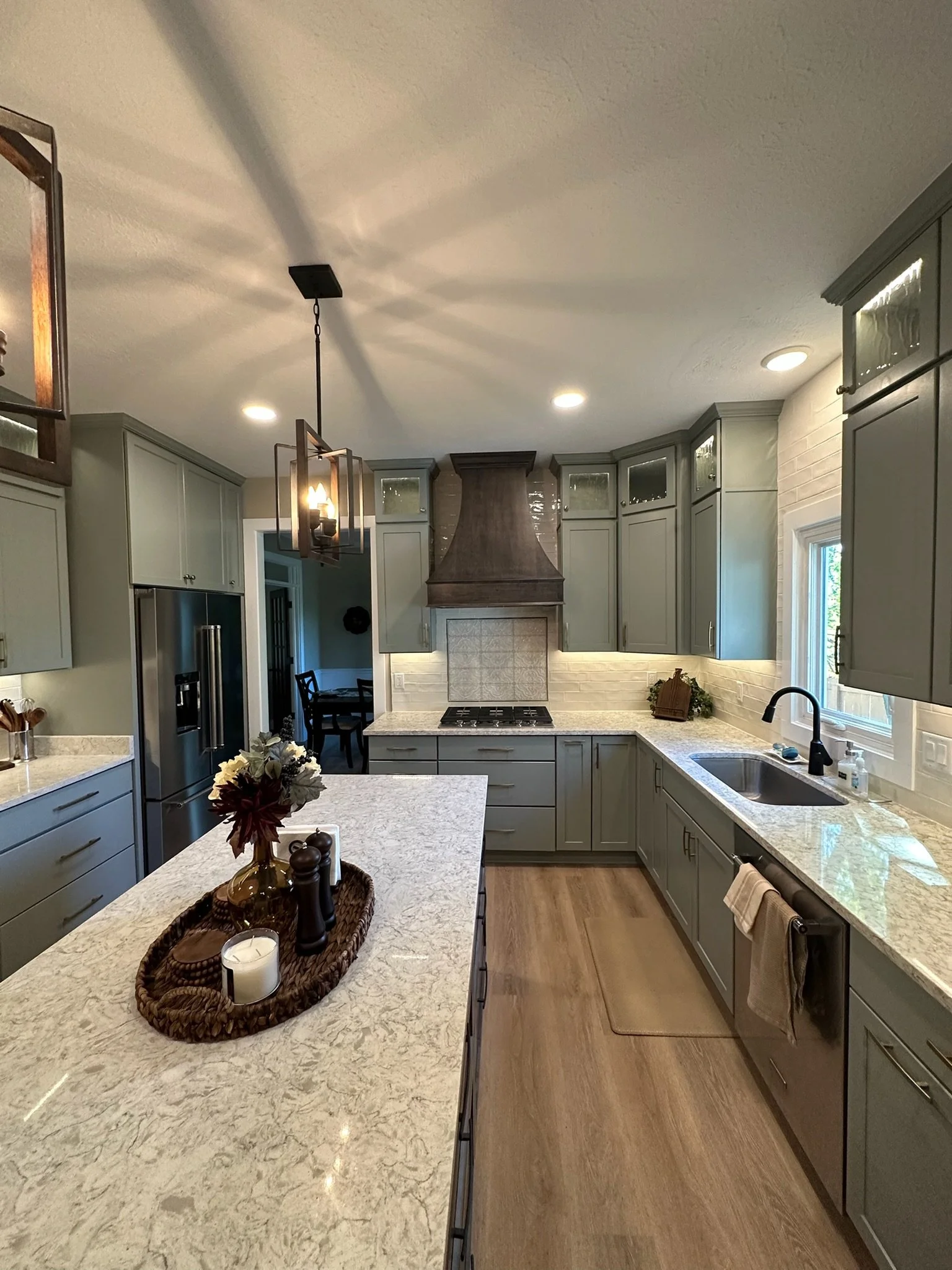 A modern kitchen with light gray cabinets, a gray stone range hood, a large window, and a white marble countertop. There is a small island with a decorative tray and candles, and a dining area with a black table and chairs in the background.