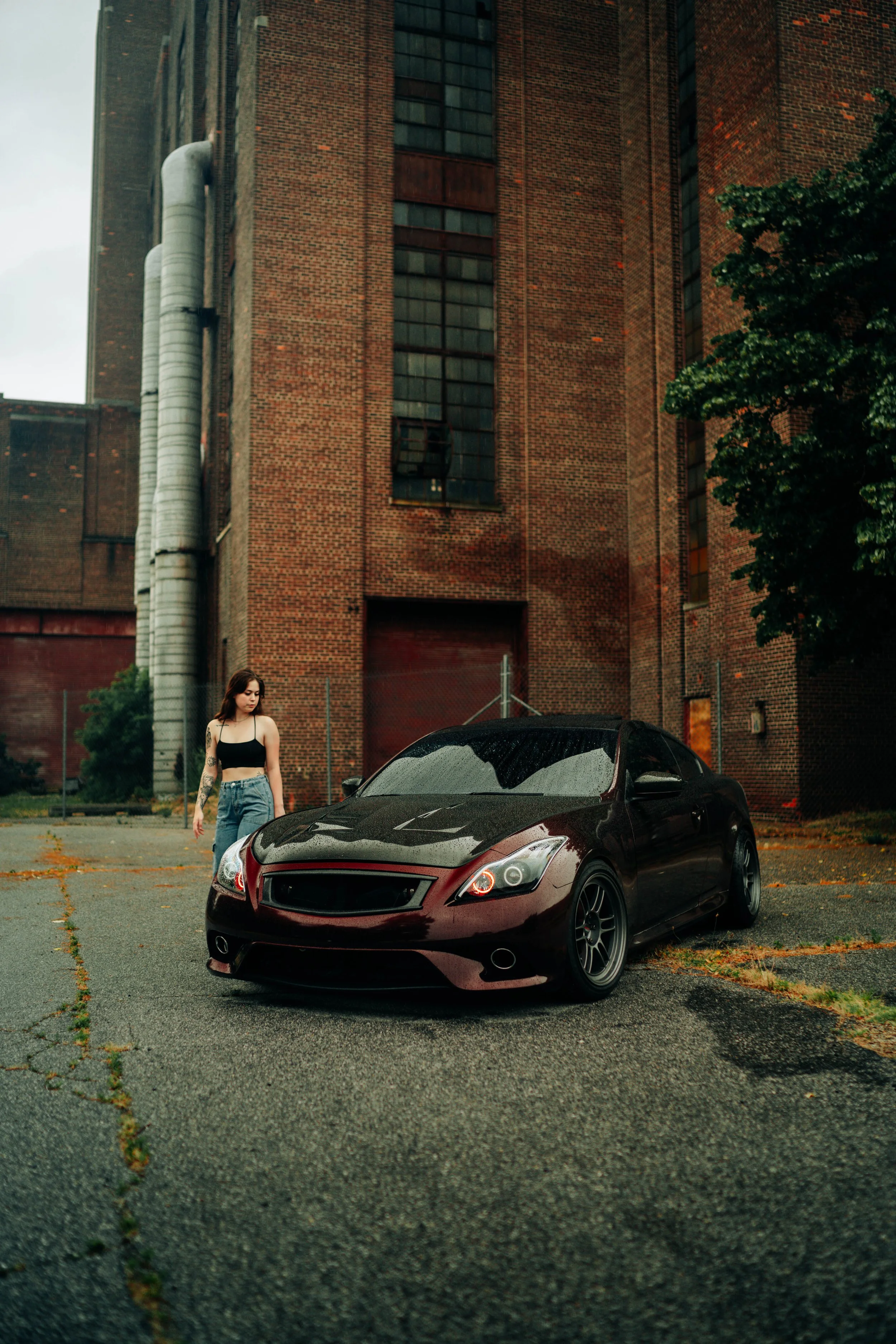 A woman with tattoos on her arm wearing a black crop top and blue jeans, standing next to a black sports car with a sleek design, in front of a brick industrial building, on an overcast day with puddles on the ground.