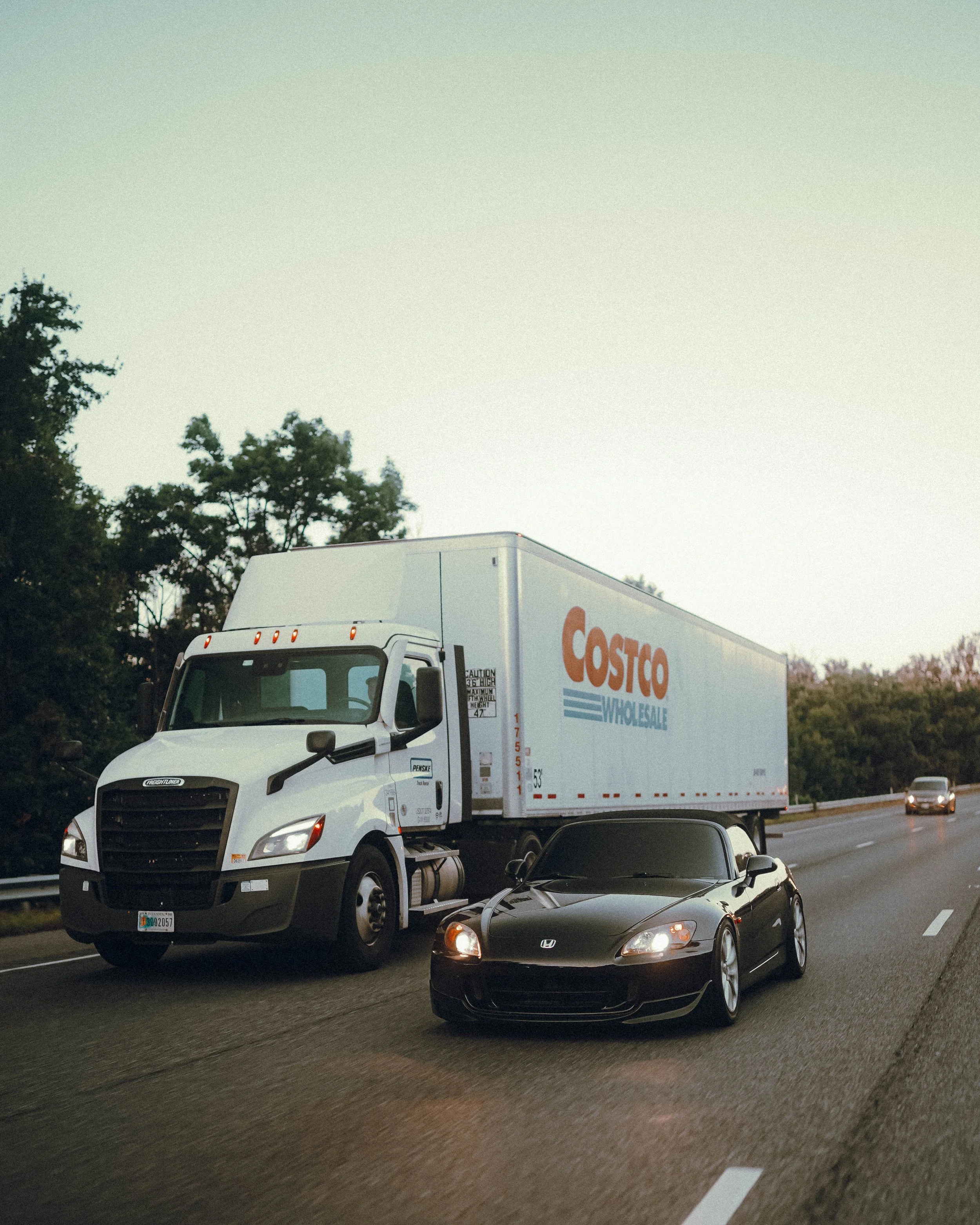 A white delivery truck with Costco Wholesale branding is traveling alongside a black car on a highway. The road is flanked by trees and other vehicles are visible in the background.