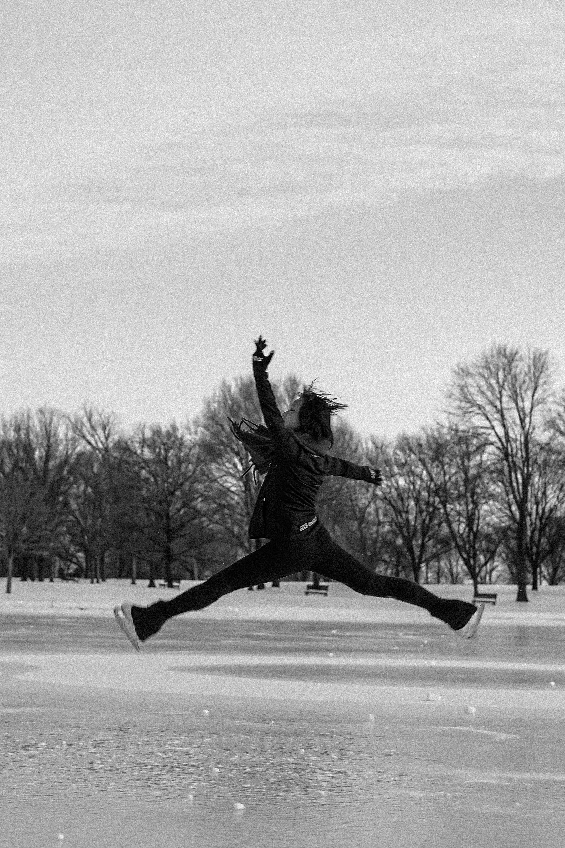 A person wearing winter clothing, including a jacket and gloves, is captured mid-air in a split leap on an icy surface during winter. The background shows leafless trees and a cloudy sky.