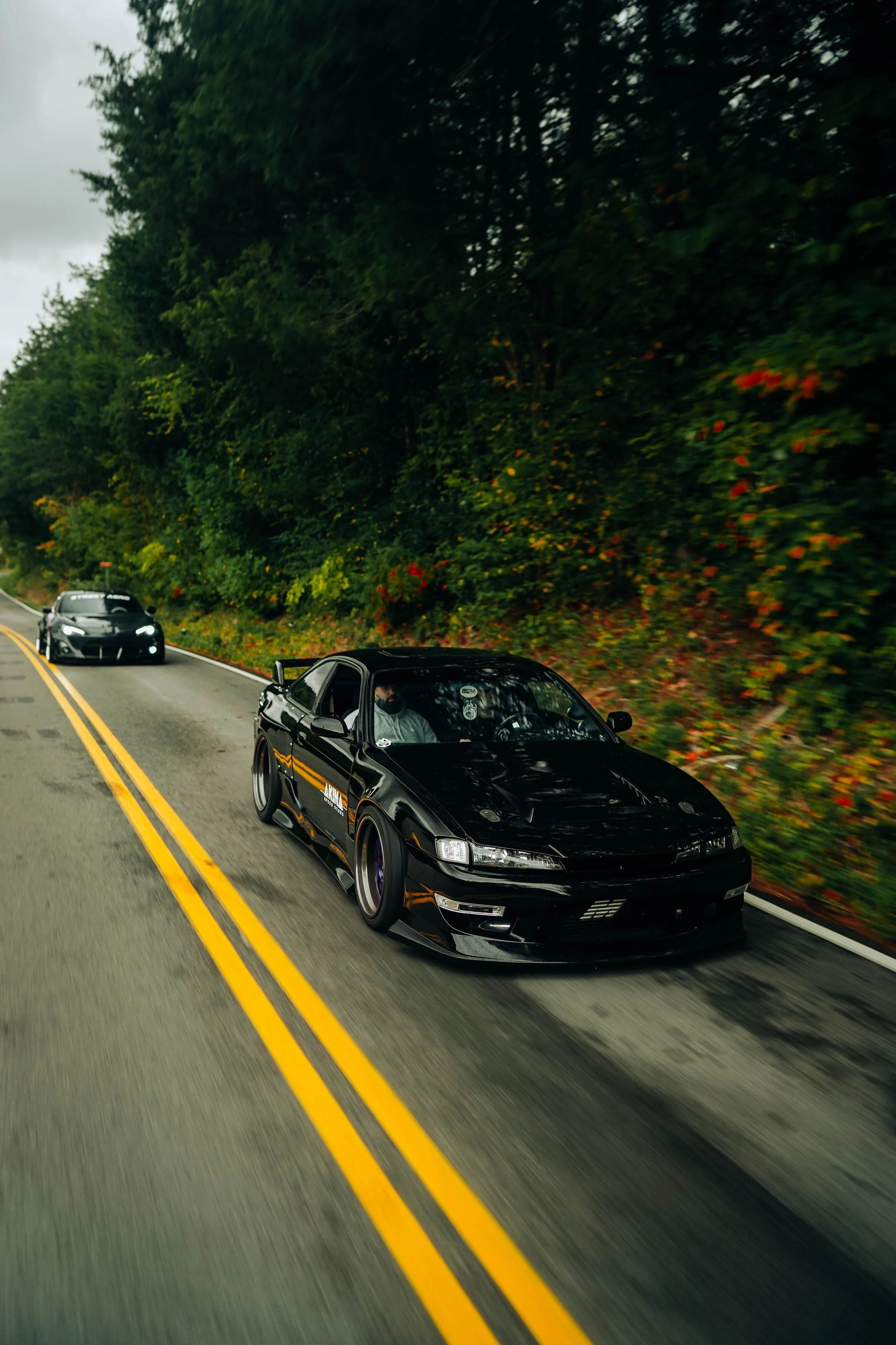 Black sports car driving on a two-lane road with a yellow center line and surrounded by dense green trees and bushes.