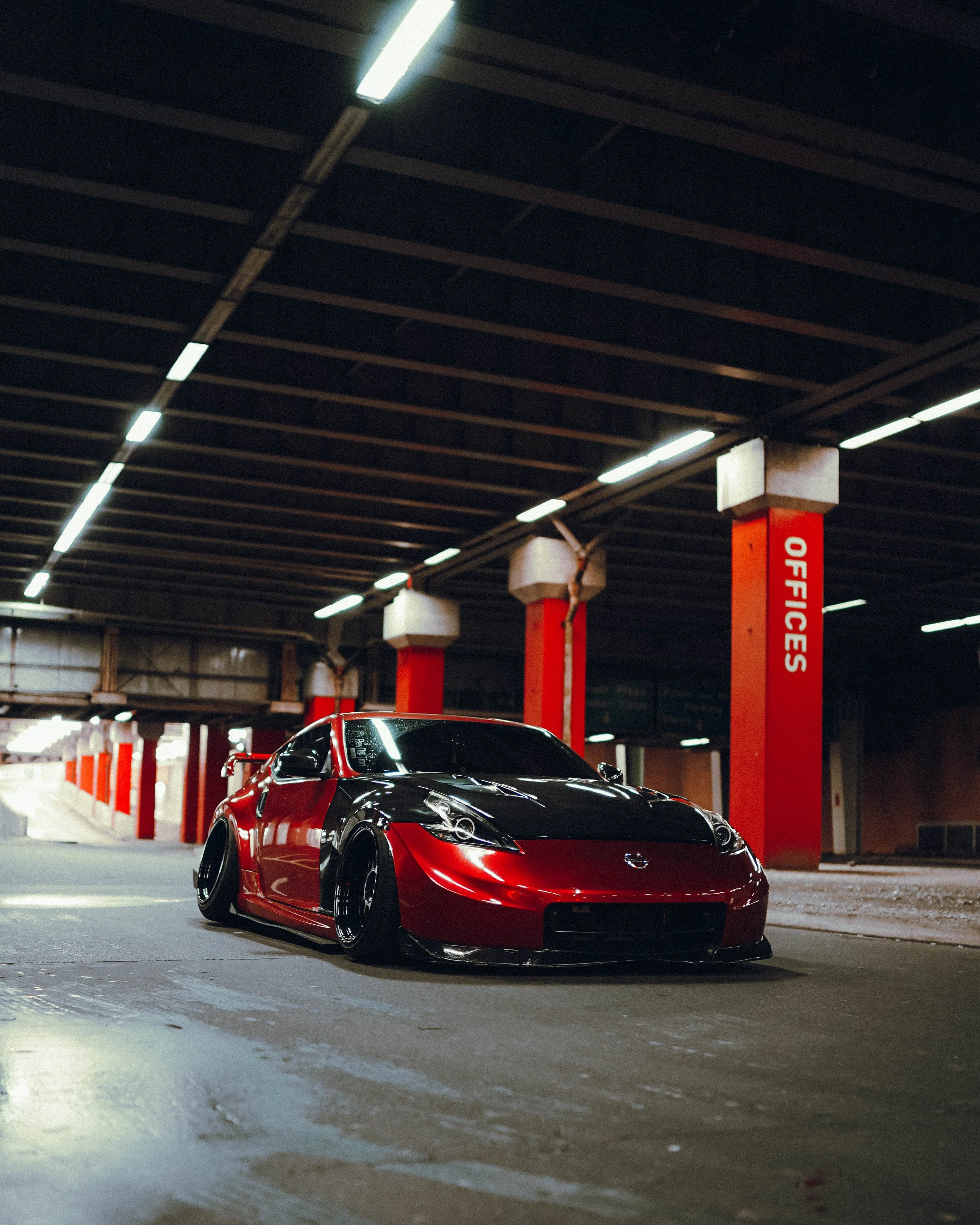A shiny red Nissan sports car with black rims is parked in an indoor parking garage with a black ceiling and red columns labeled 'OFFICES'.