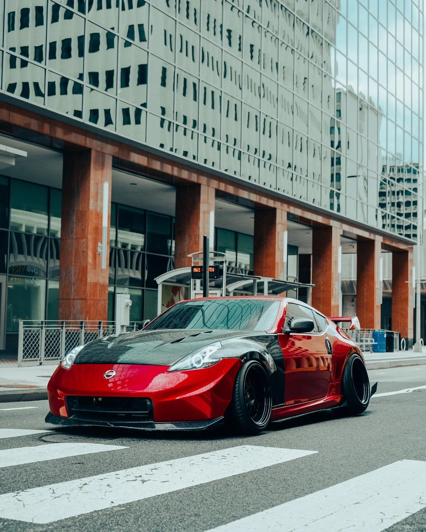 A red and black sports car parked on the street in front of a modern glass building with reflections of other buildings.