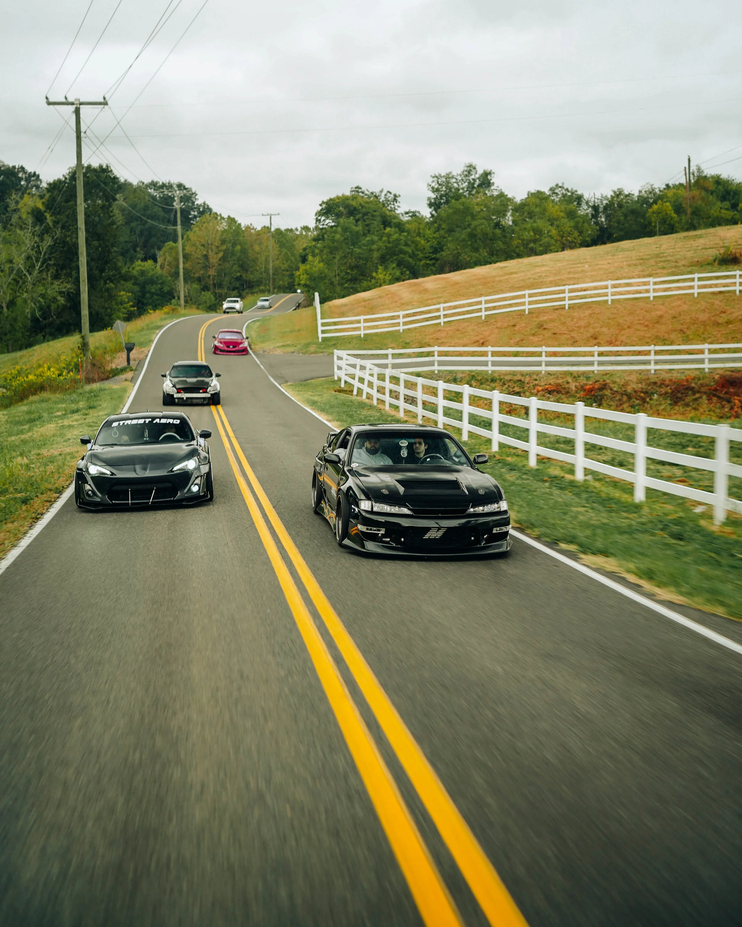 A winding rural road with cars traveling in both directions, flanked by grassy areas, white fences, and trees, under an overcast sky.