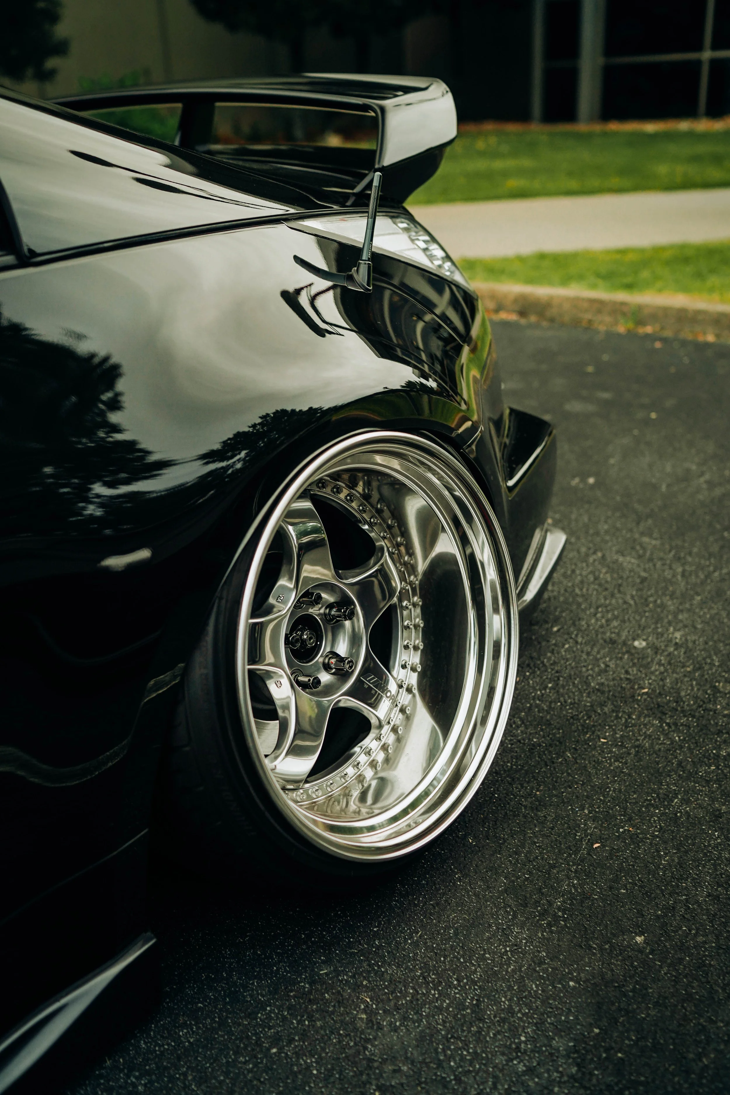 Close-up of a black sports car with a custom wheel and tire, showing part of the wheel well, rear spoiler, and antenna, parked on a dark asphalt driveway with a grassy area and a building in the background.