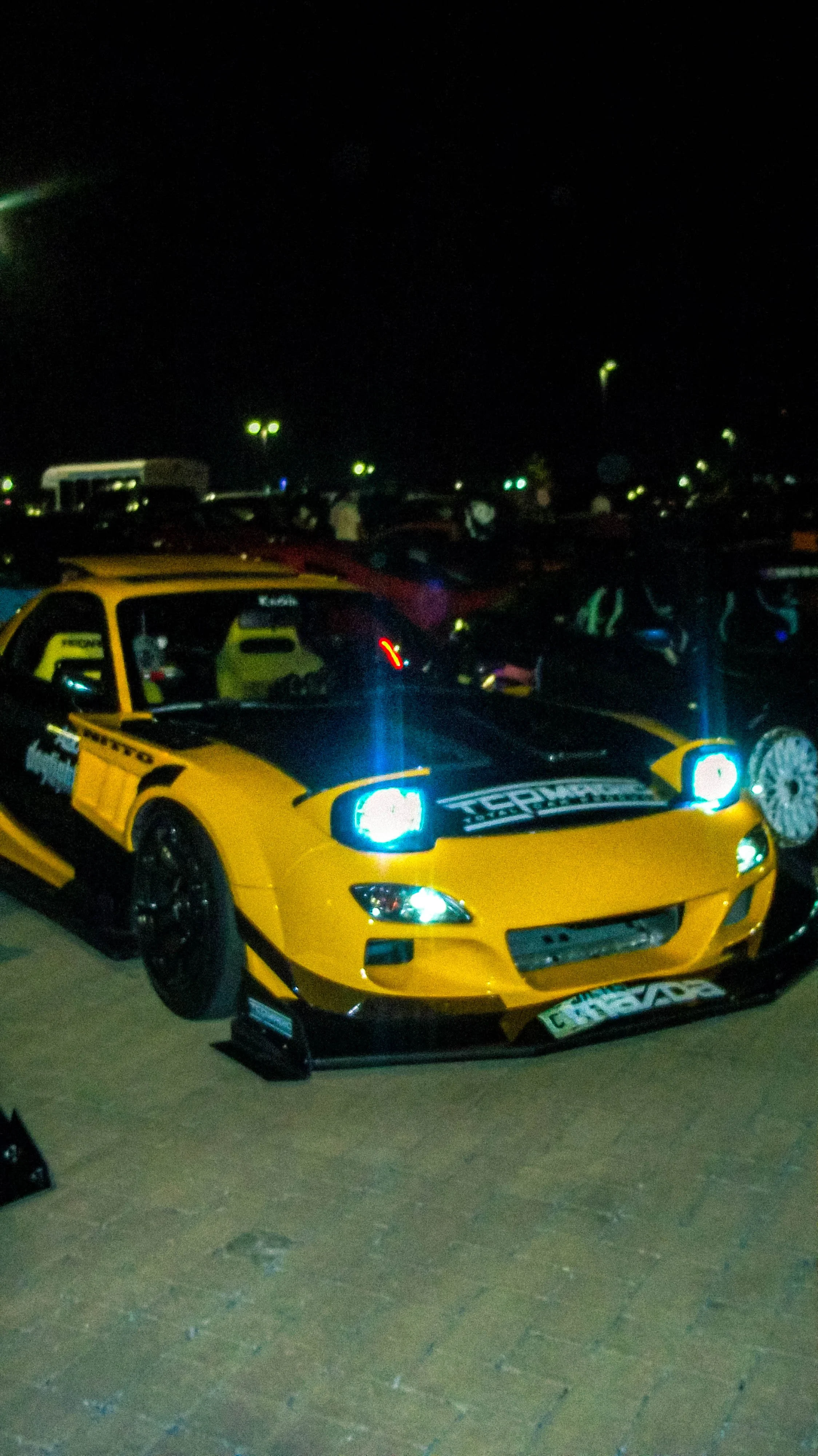 A yellow sports car parked on pavement at night, with bright headlights and racing decals, among other cars in a dimly lit lot.