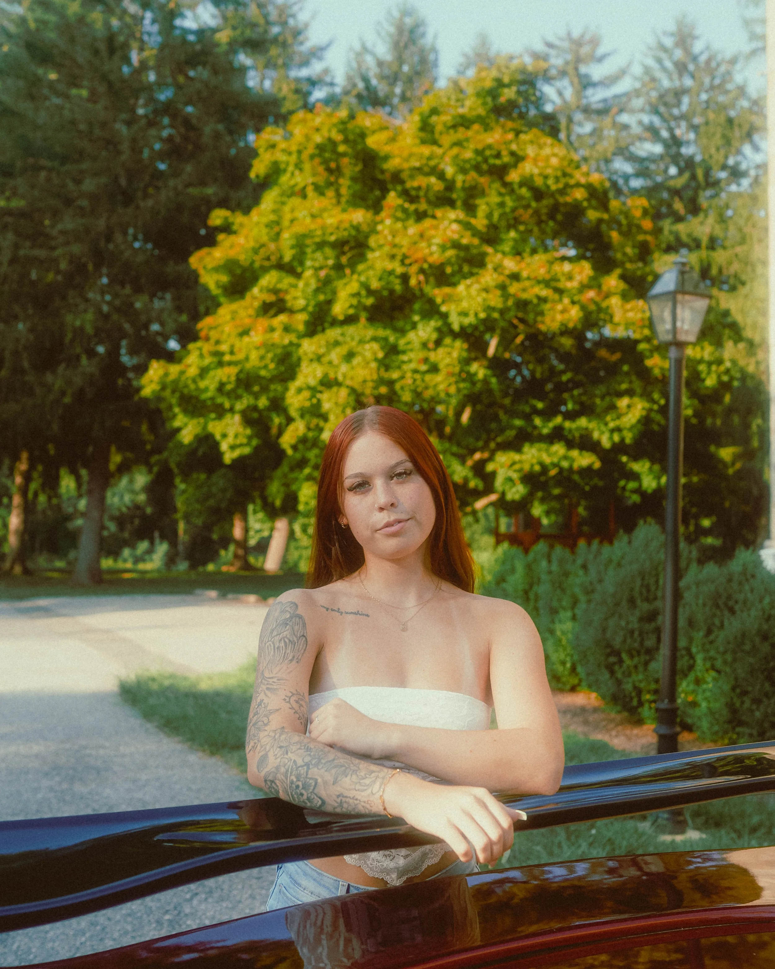 A young woman with long red hair standing outdoors in front of trees with green and yellow leaves, crossing her arms, and leaning on the roof of a black vehicle.