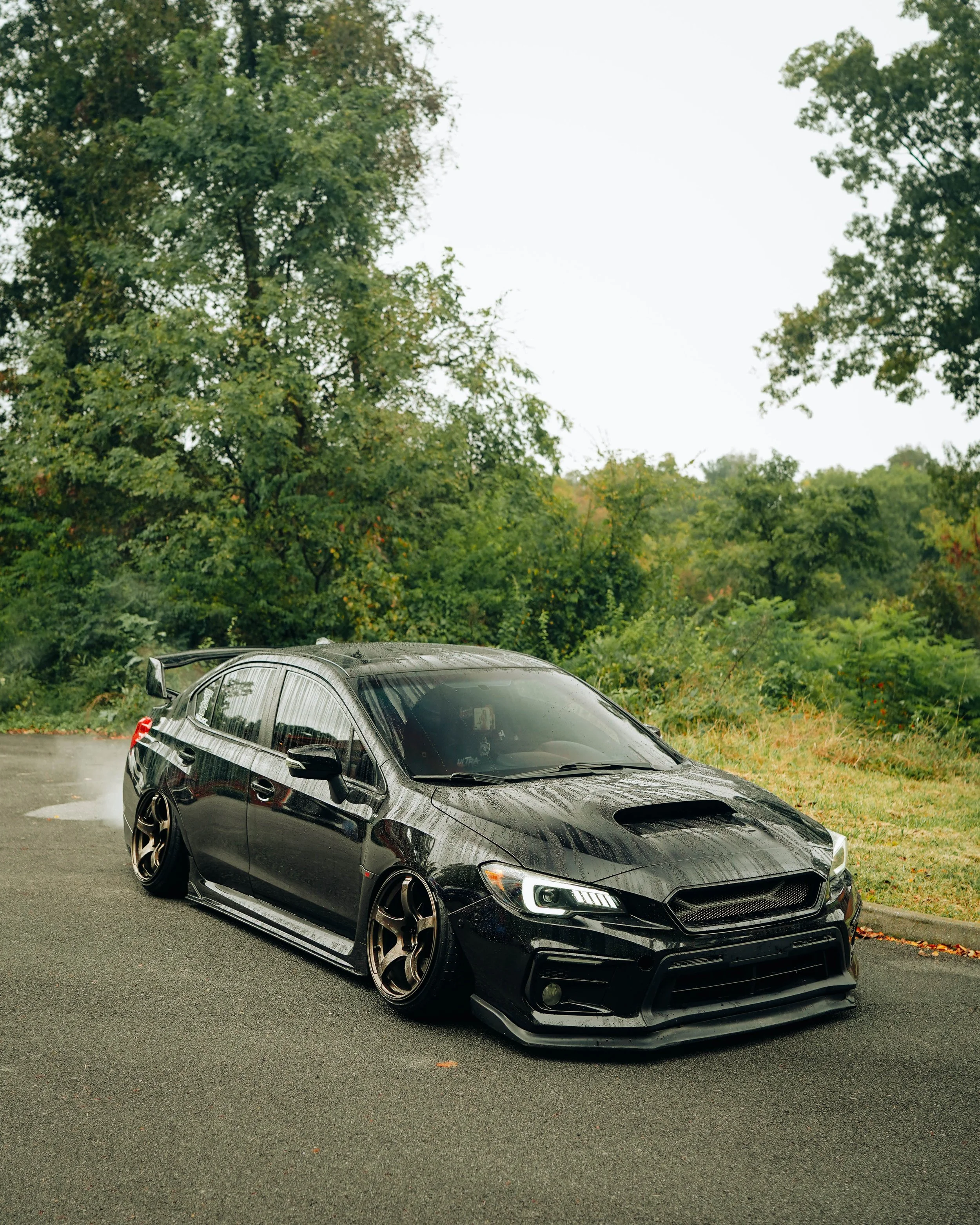 A black sports car with a custom wide body kit parked on a paved lot, with green trees and bushes in the background and an overcast sky.