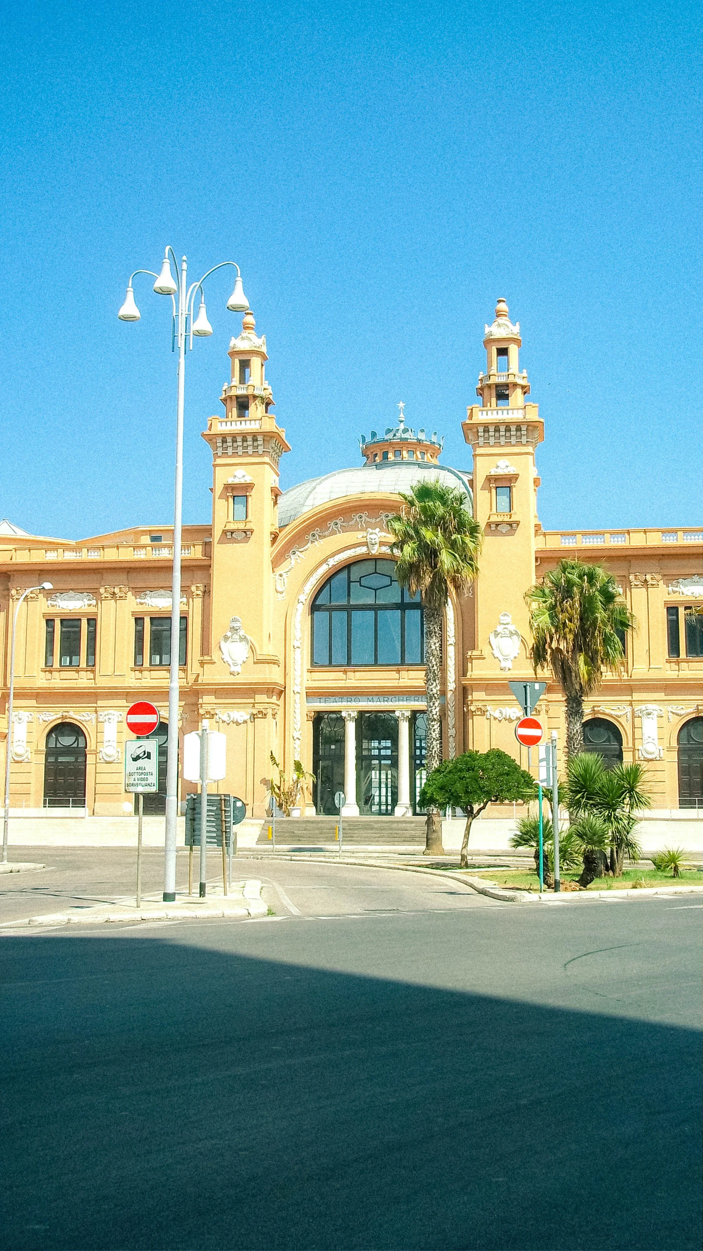 Front view of an ornate yellow building with tall towers and a large arched entrance, flanked by some palm trees, under a clear blue sky.