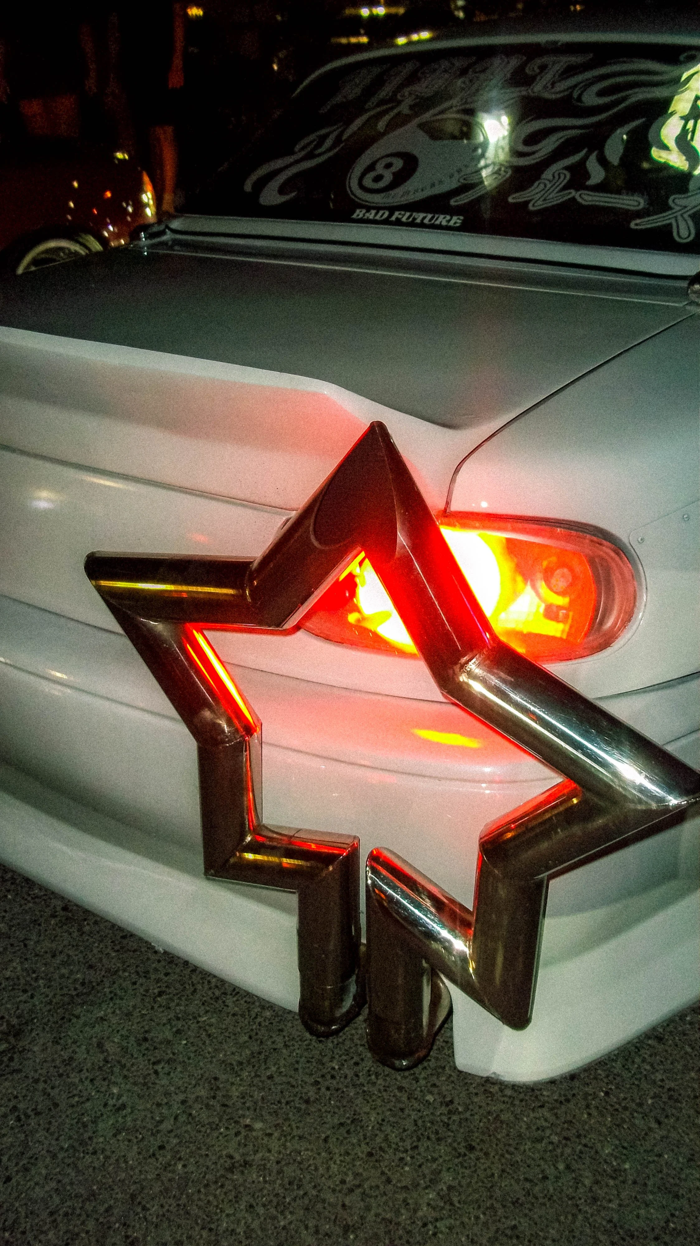 Close-up of the rear of a white car with a chrome star emblem on the trunk. The car has a taillight illuminated and a sticker on the rear windshield that reads "BAD FUTURE" above a billiard ball with the number 8. There is a prominent chrome star-sha