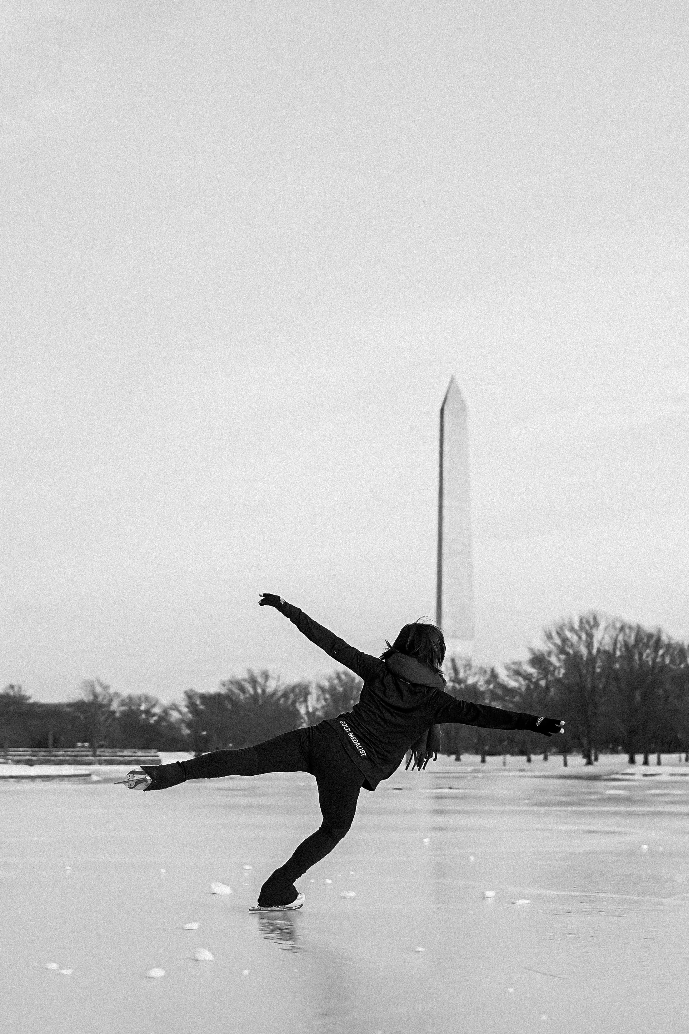A person ice skating outdoors on a frozen lake during the winter, performing a professional move with arms outstretched and one leg extended behind, in front of a background of trees and the Washington Monument.