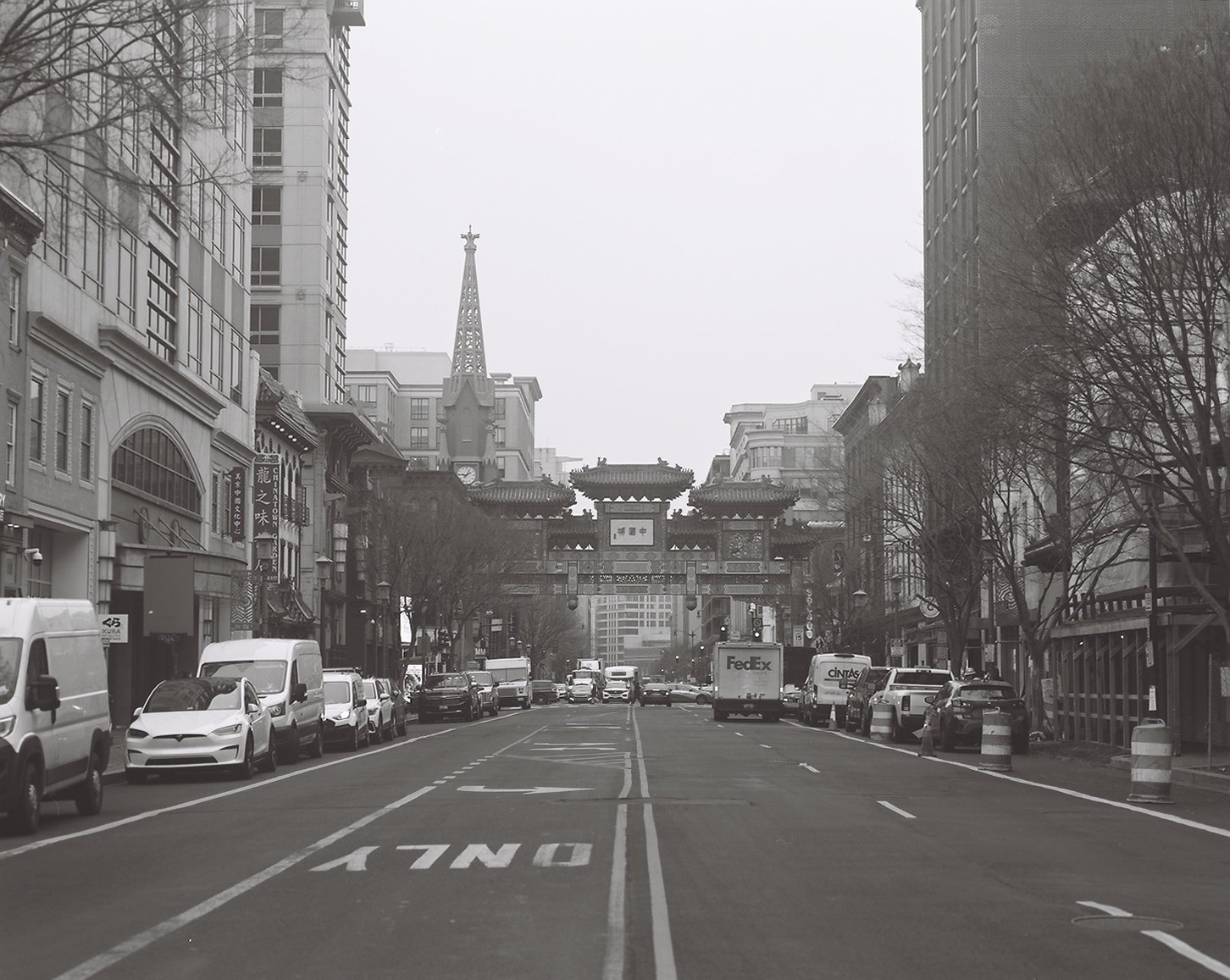A city street scene in grayscale with parked cars on both sides and a traditional Chinese-style gate in the distance, with modern buildings and a church steeple in the background.