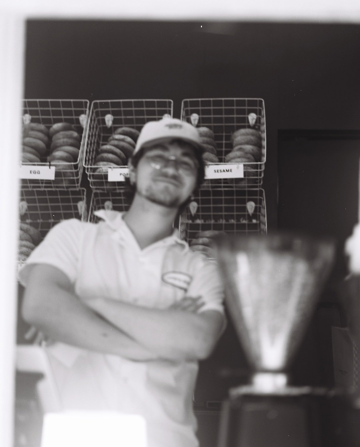 A man wearing a cap and white shirt standing in front of wire baskets filled with baked goods labeled 'Egg,' 'Potato,' and 'Sesame' in a bakery or store.