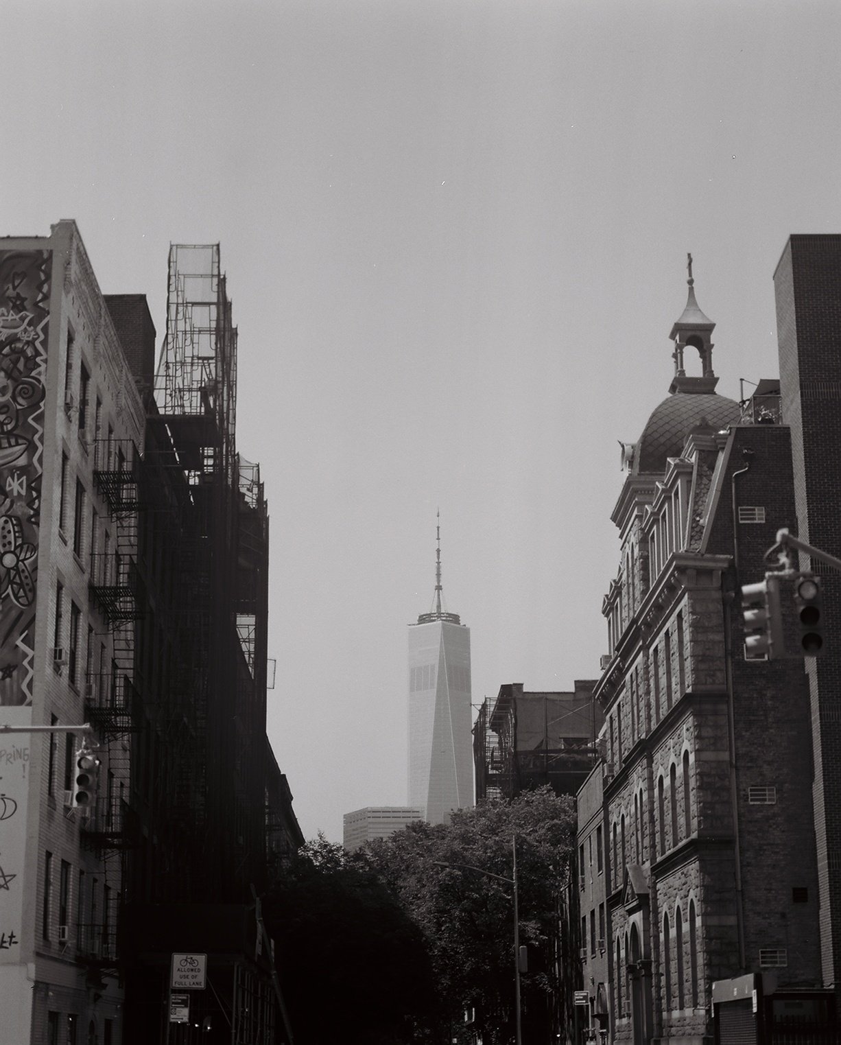 Black and white photo of a city street with buildings on each side, One World Trade Center in the background, and a historic building with a dome and a spire on the right.