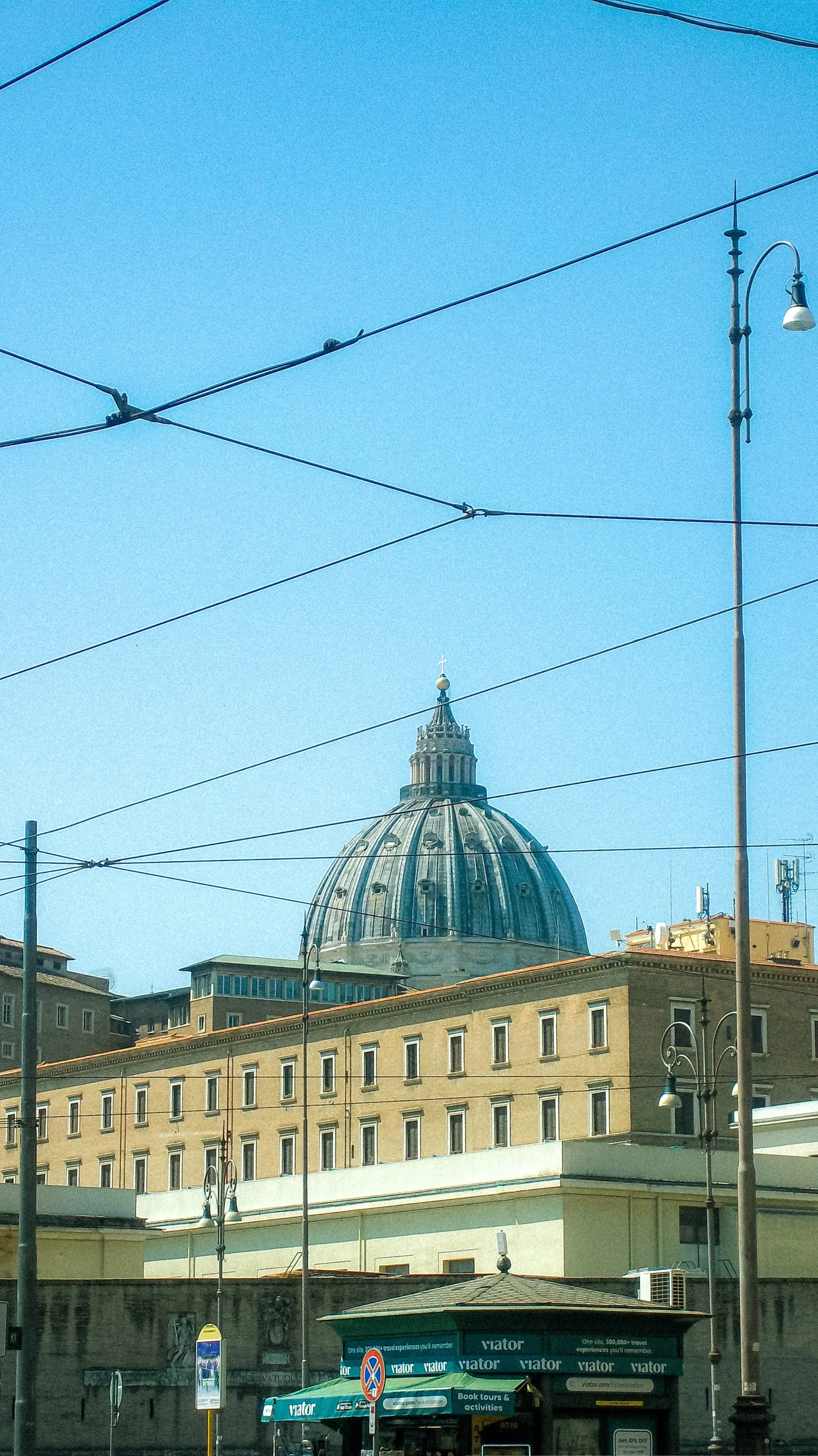 Cityscape with the dome of St. Peter's Basilica in Vatican City, Rome, Italy, visible in the background against a clear blue sky. Overhead tram wires and street lamps are in the foreground, along with buildings and a shop with a sign that reads 'vato