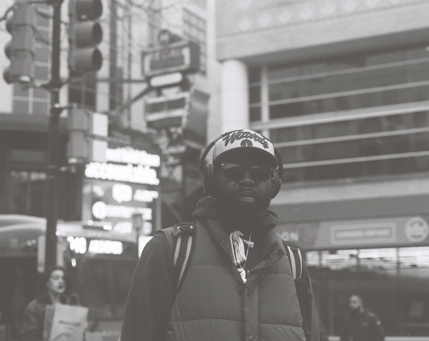 A man wearing sunglasses, a hat that says 'Wizards', and a jacket with a backpack, standing on a city street with blurred buildings and people in the background.