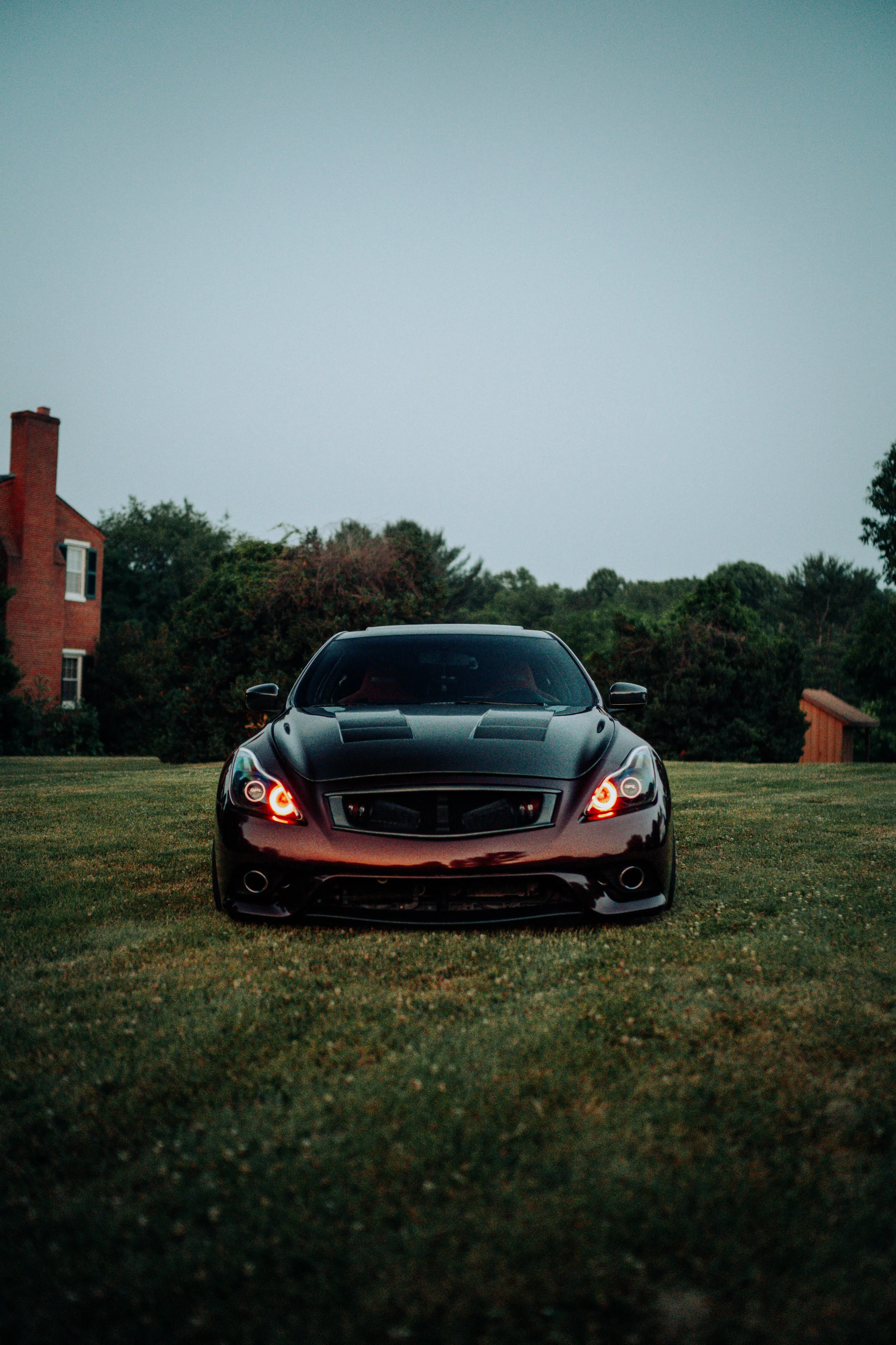 Front view of a black sports car parked on a grassy area with trees and a brick house in the background, under an overcast sky.