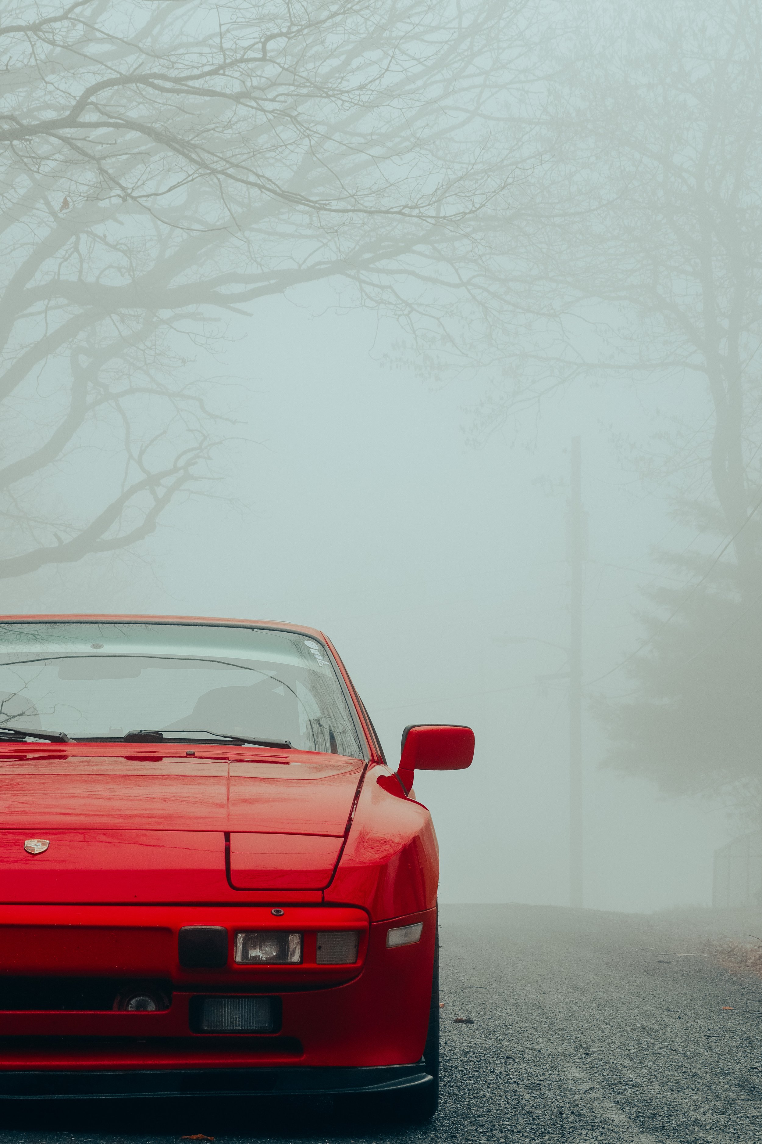 A red Porsche 944 sports car parked on a street with foggy, leafless trees in the background. The front of the car is prominently displayed, showing the hood, windshield, and side mirror.
