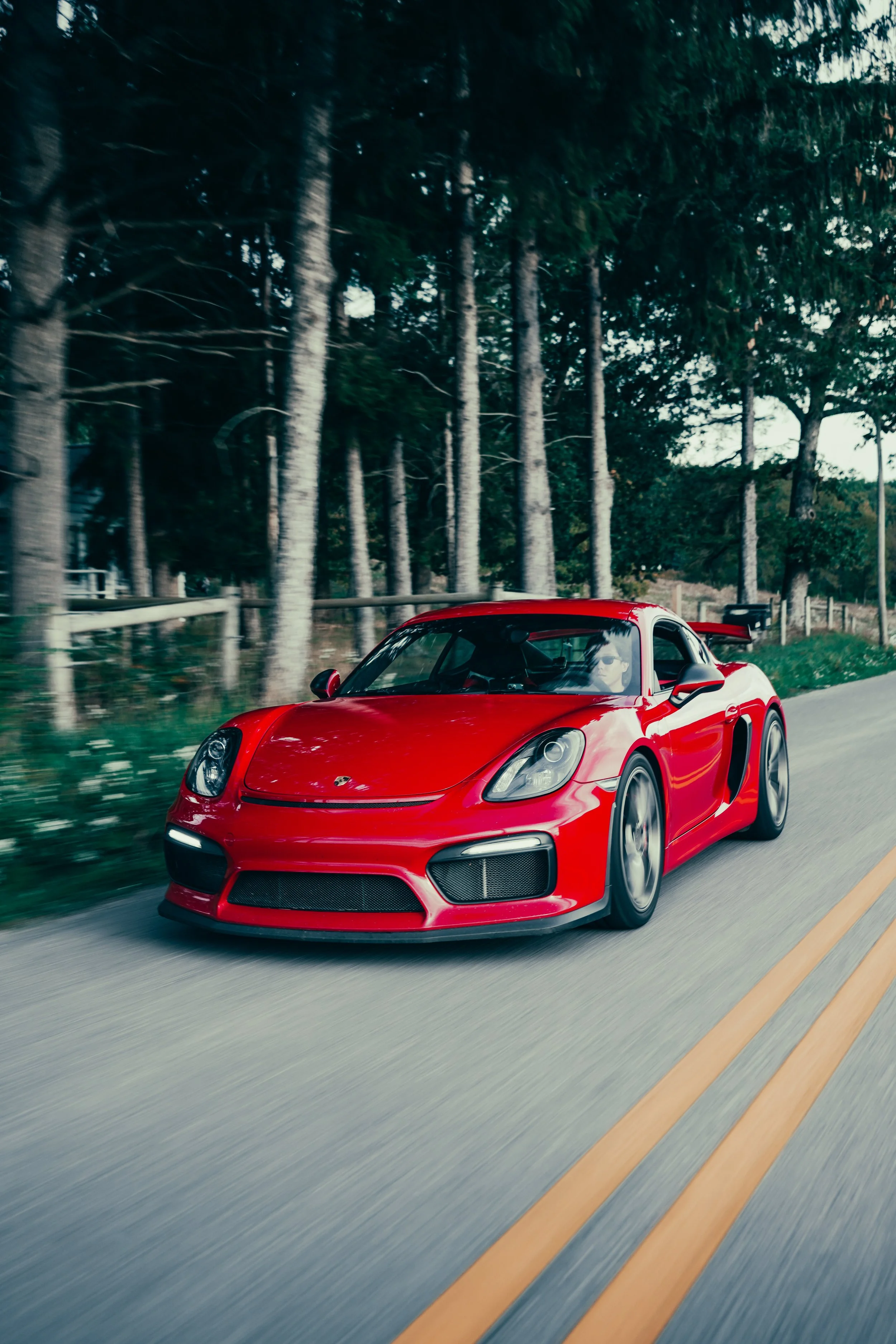 A red sports car driving on a rural road with trees and a wooden fence in the background.