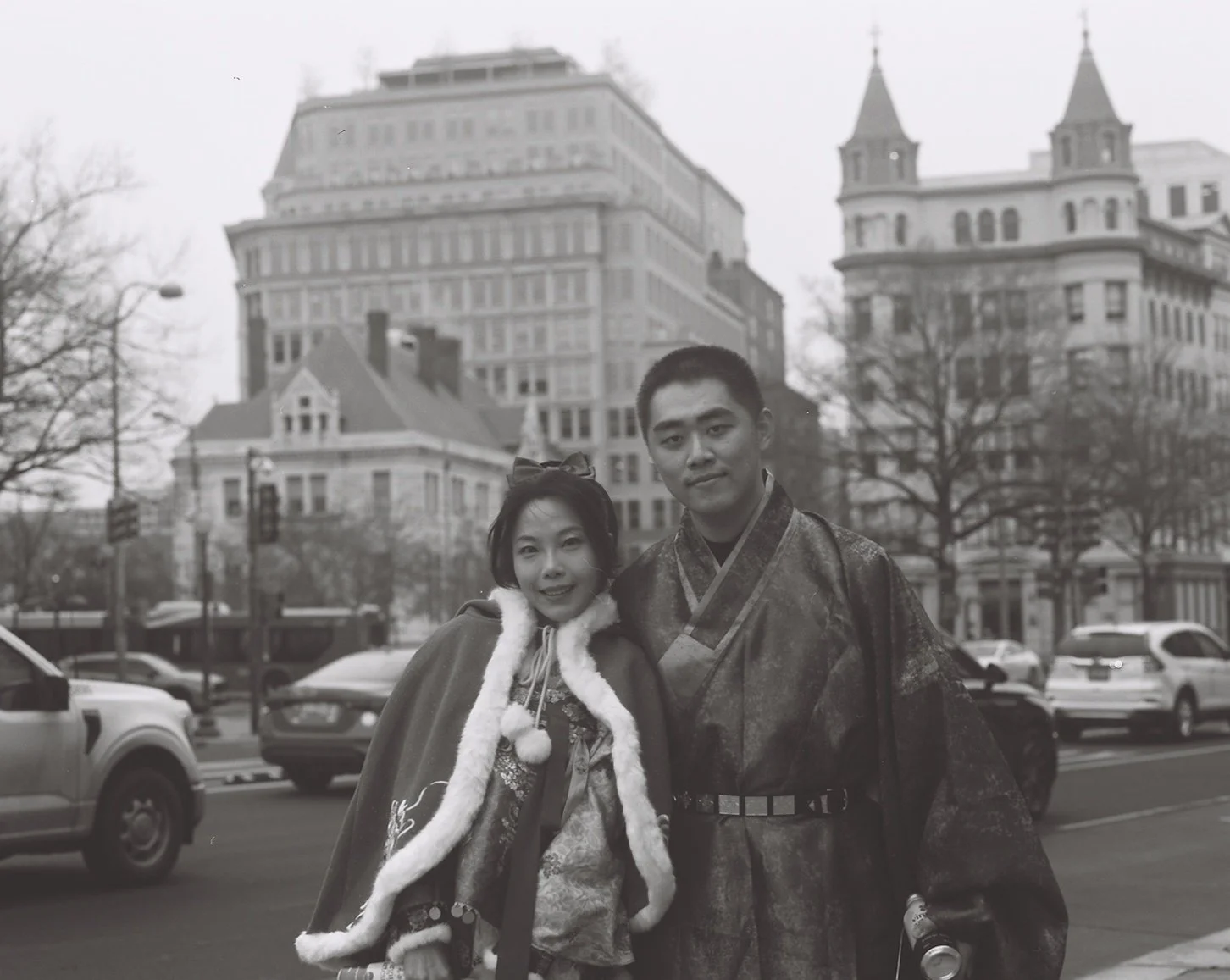 A black and white photo of a woman and a man in traditional Asian clothing standing on a city street. There are cars parked and in motion behind them, and large historic buildings with trees in the background.