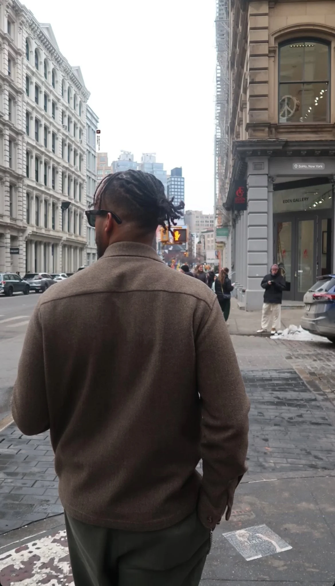 A man with dreadlocks, sunglasses, and a tan coat stands on a city sidewalk with tall buildings, people, cars, and a crosswalk in the background, in SoHo, New York.