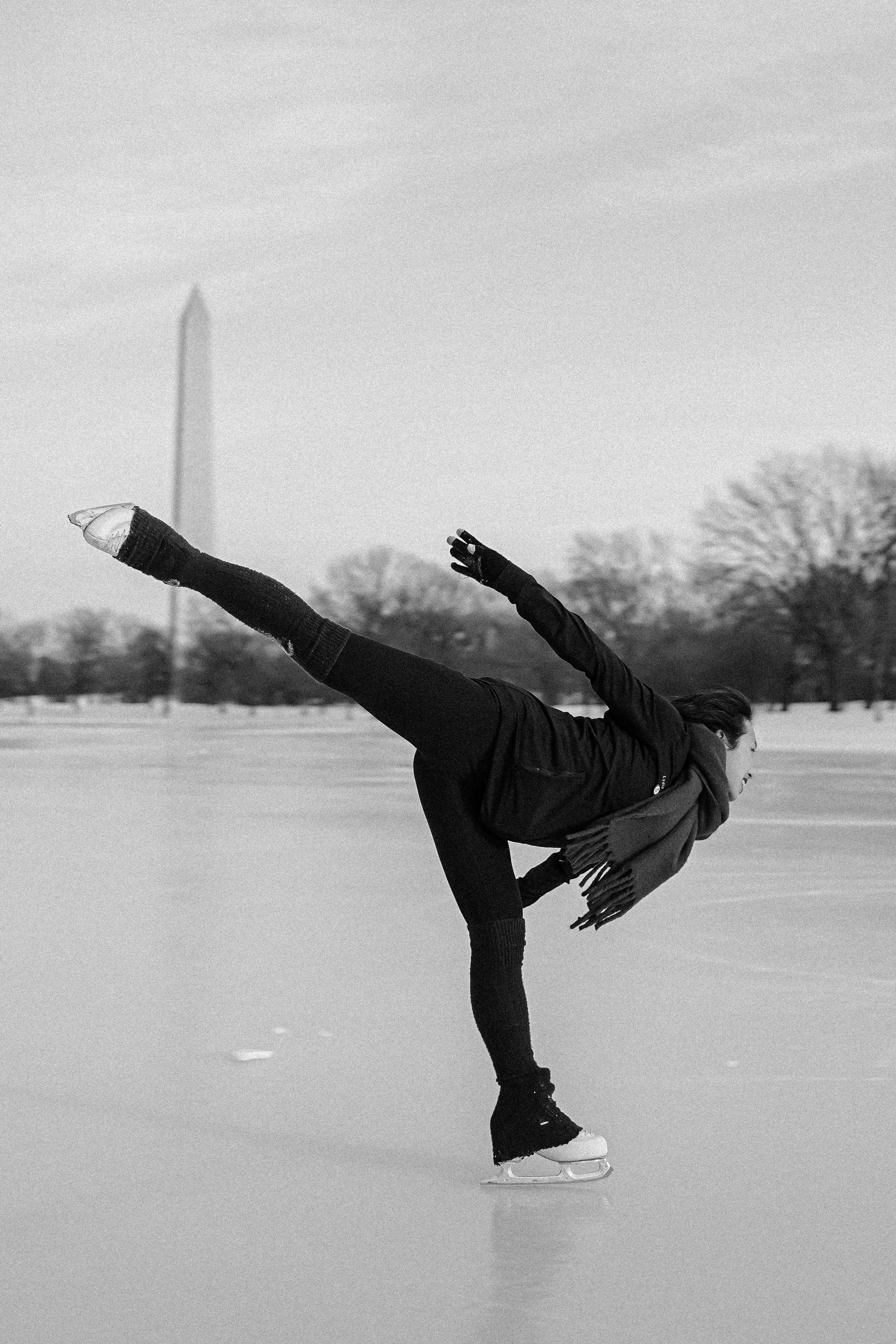 Person skating on ice, performing a one-legged balancing move, outdoors in winter, with trees in the background and the Washington Monument in the distance.